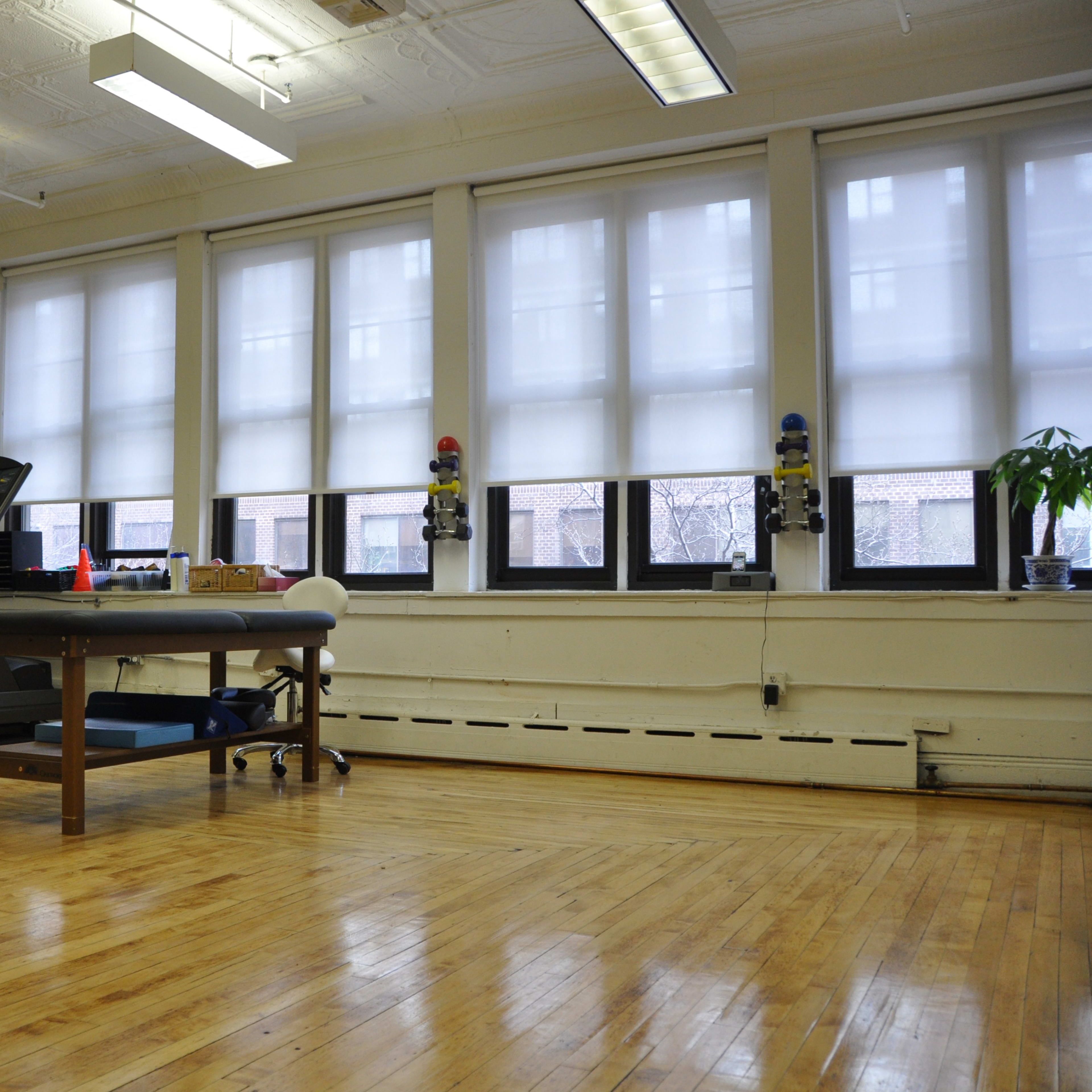 A spacious room with wooden floors, featuring a treadmill, a table, and large windows with blinds.