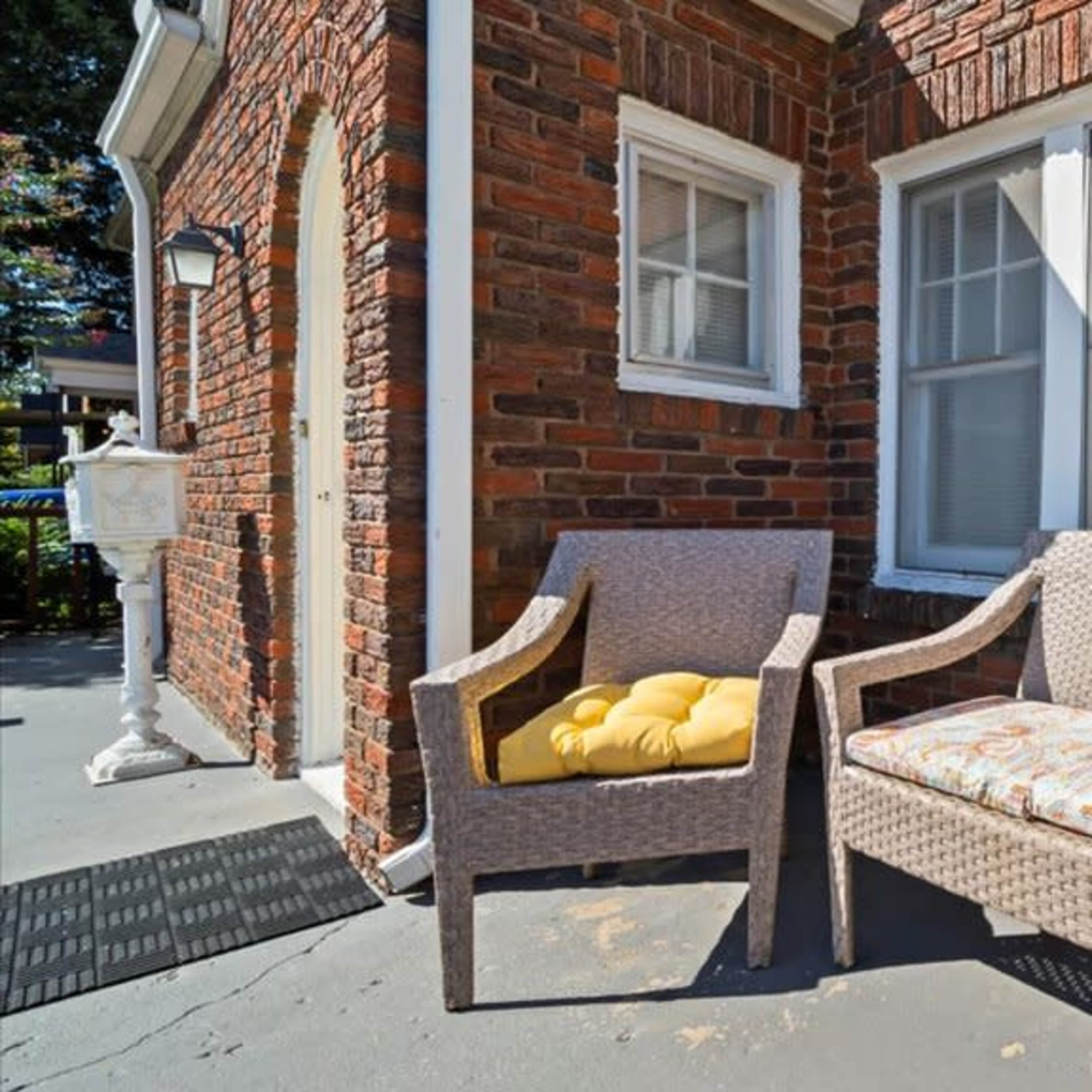 A small outdoor seating area with two wicker chairs and decorative cushions beside a brick exterior wall.