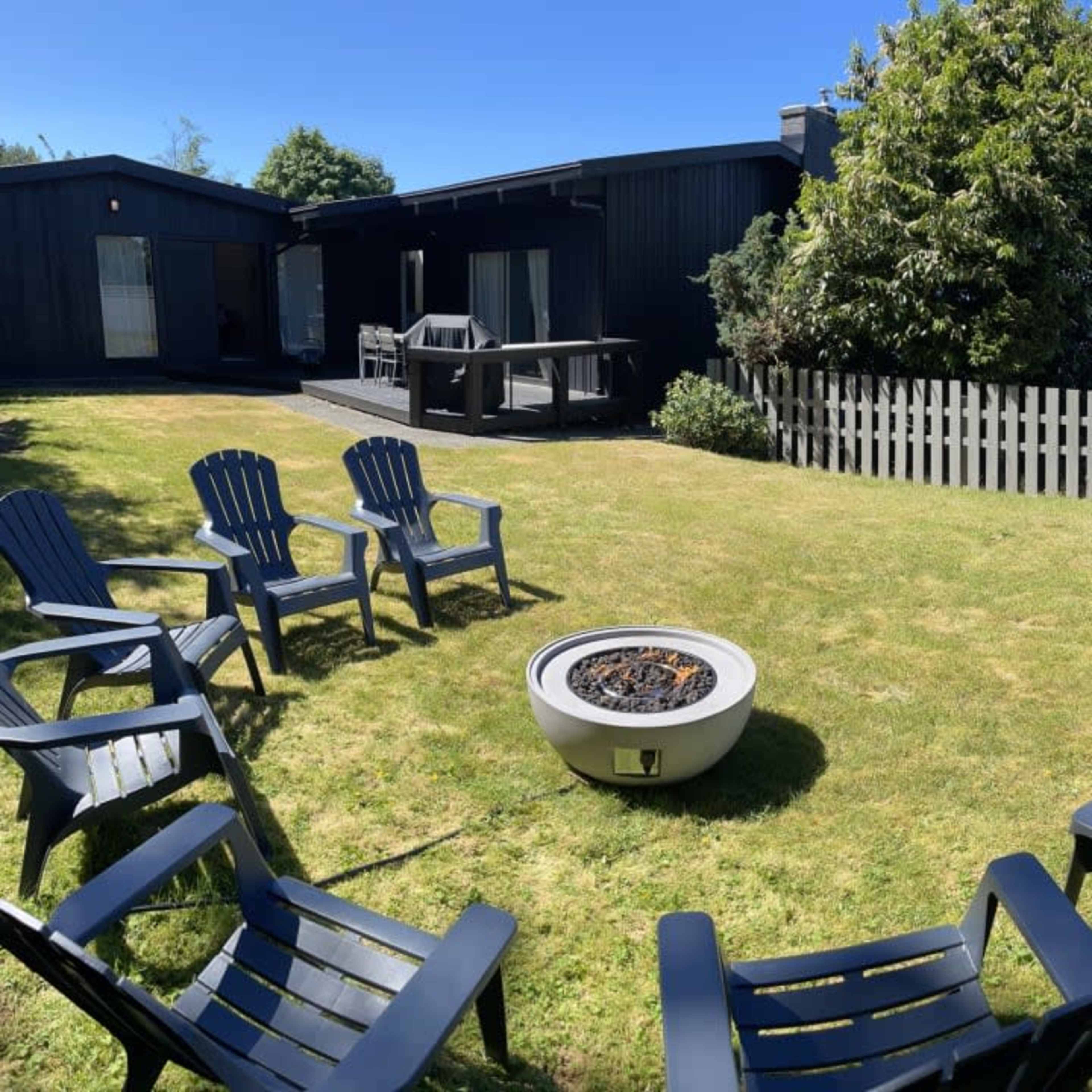 A circular arrangement of eight black chairs surrounds a fire pit in a grassy yard overlooking the ocean.