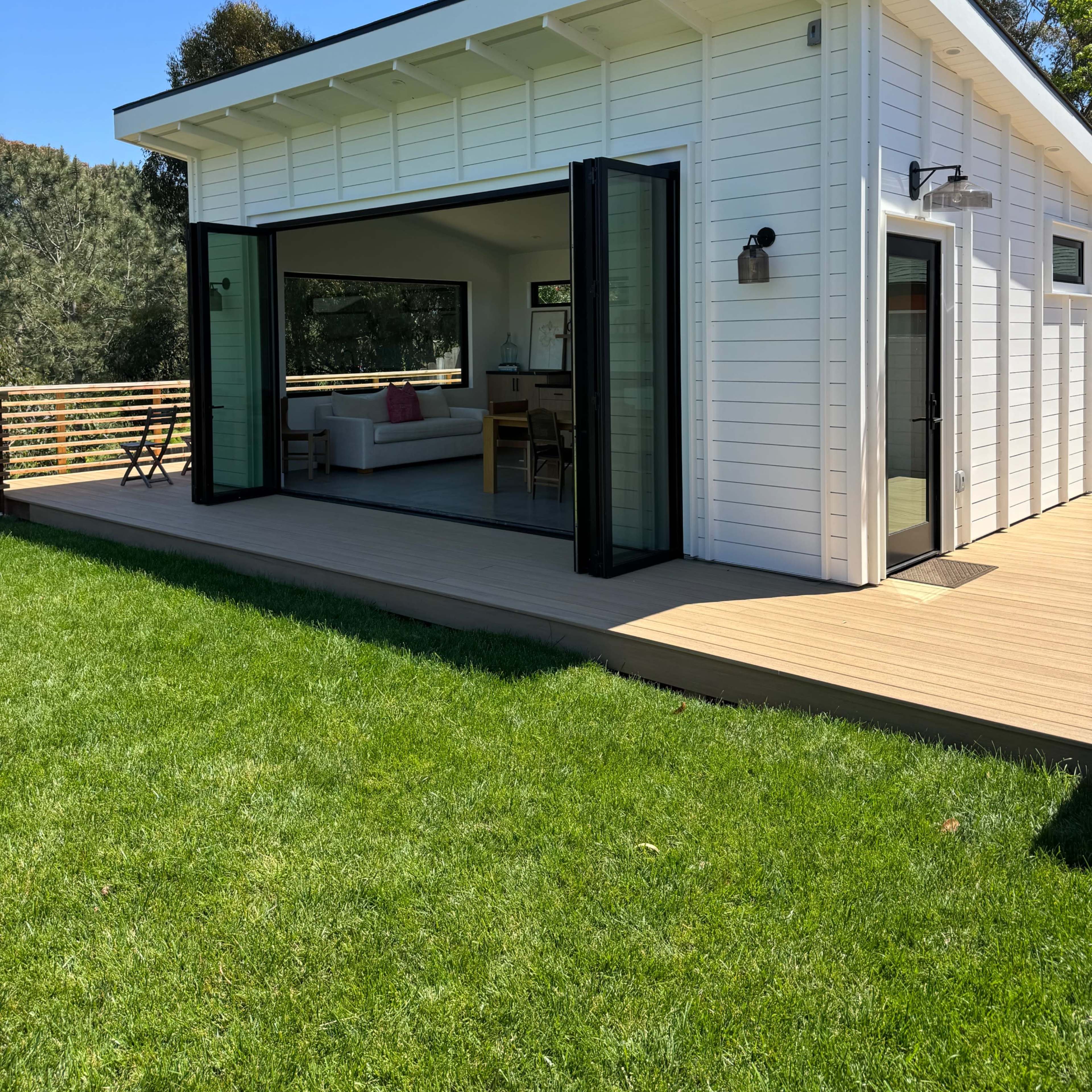 A modern, white-clad building features large glass doors that open onto a wooden deck, overlooking a green lawn and surrounded by trees.