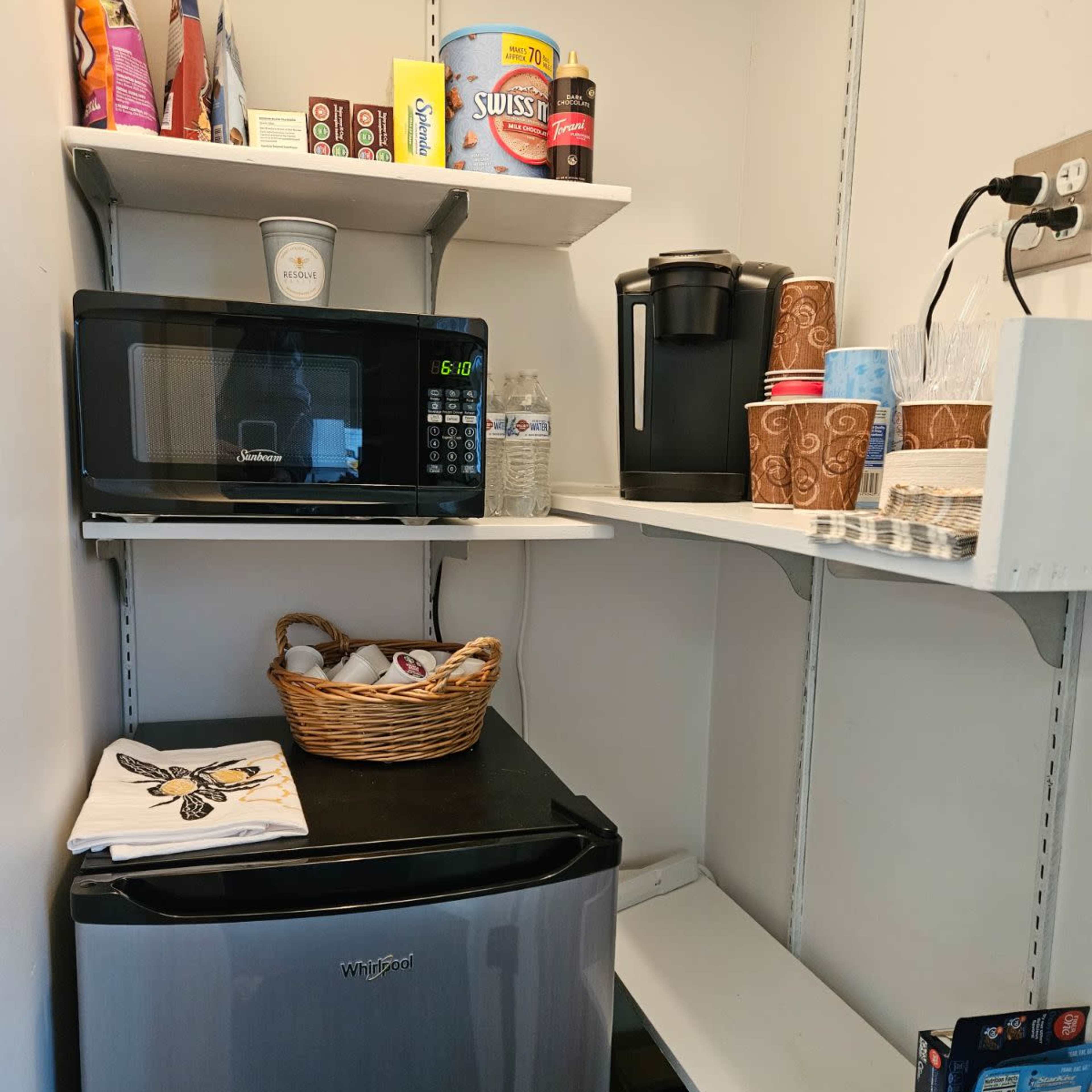 A small kitchenette area with a mini fridge, microwave, coffee maker, and organized shelves containing cups and snacks.