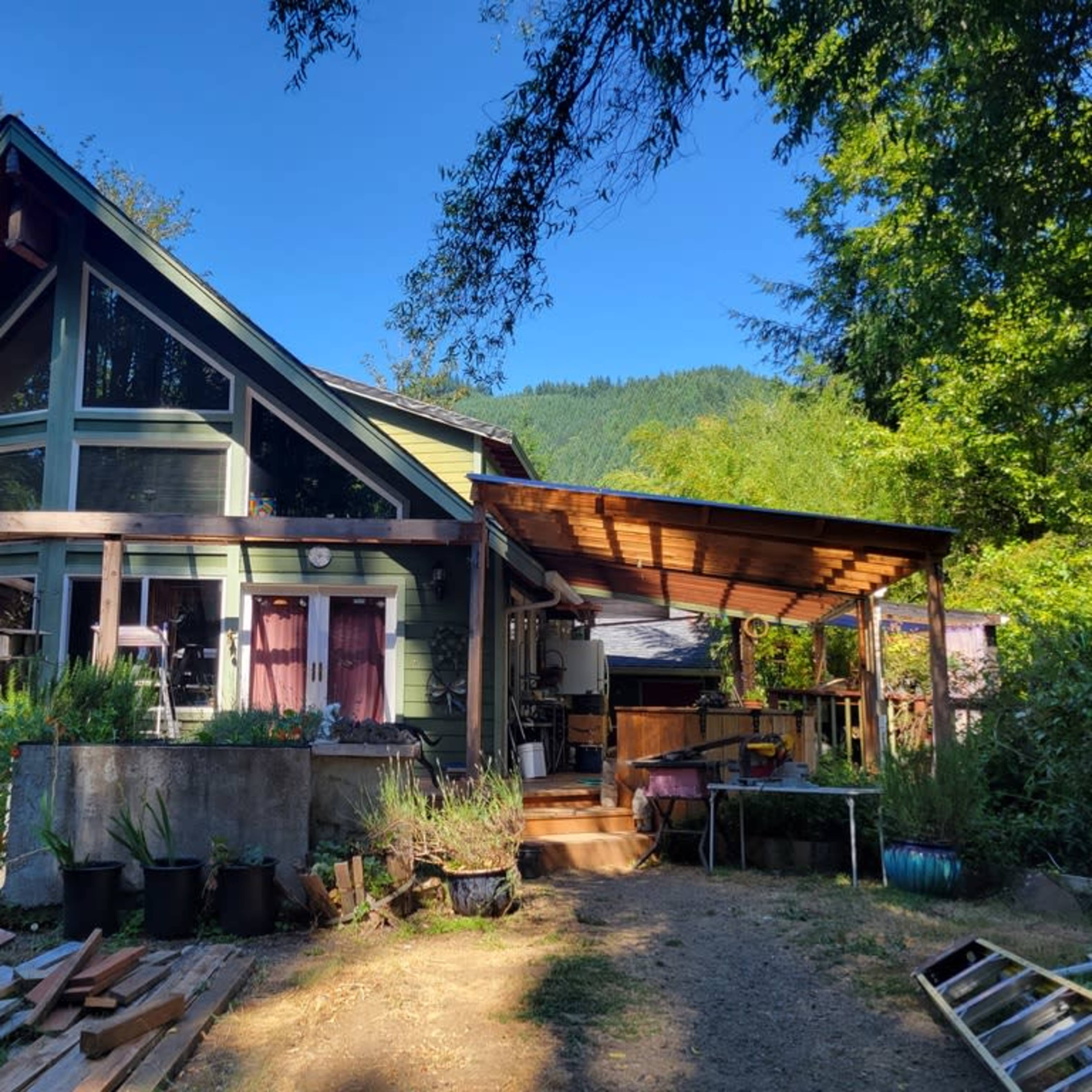 A green and brown house with a large deck, surrounded by trees and gardening supplies.