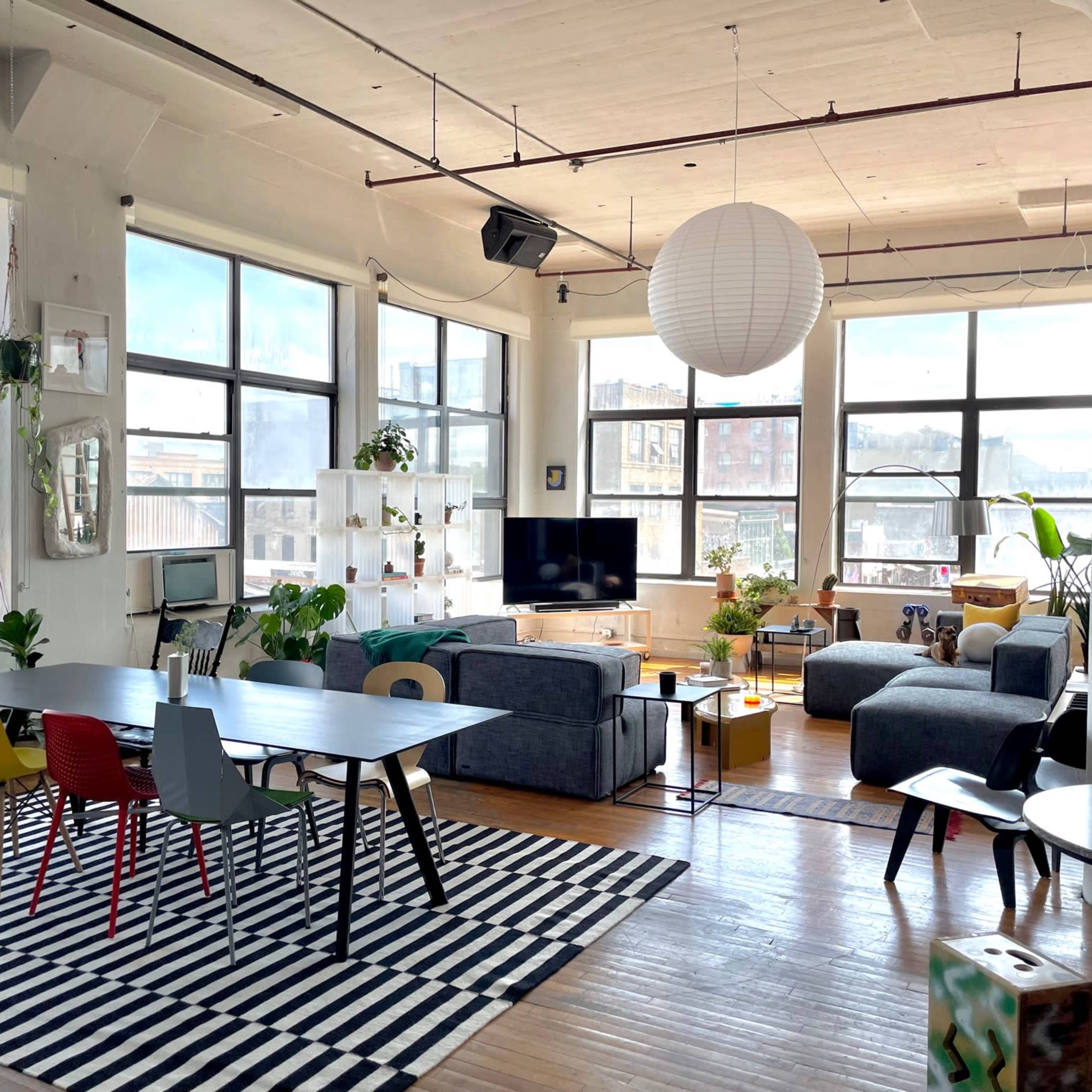 A bright, open living space with a large window, featuring a mix of seating areas, a dining table, and decorative plants.