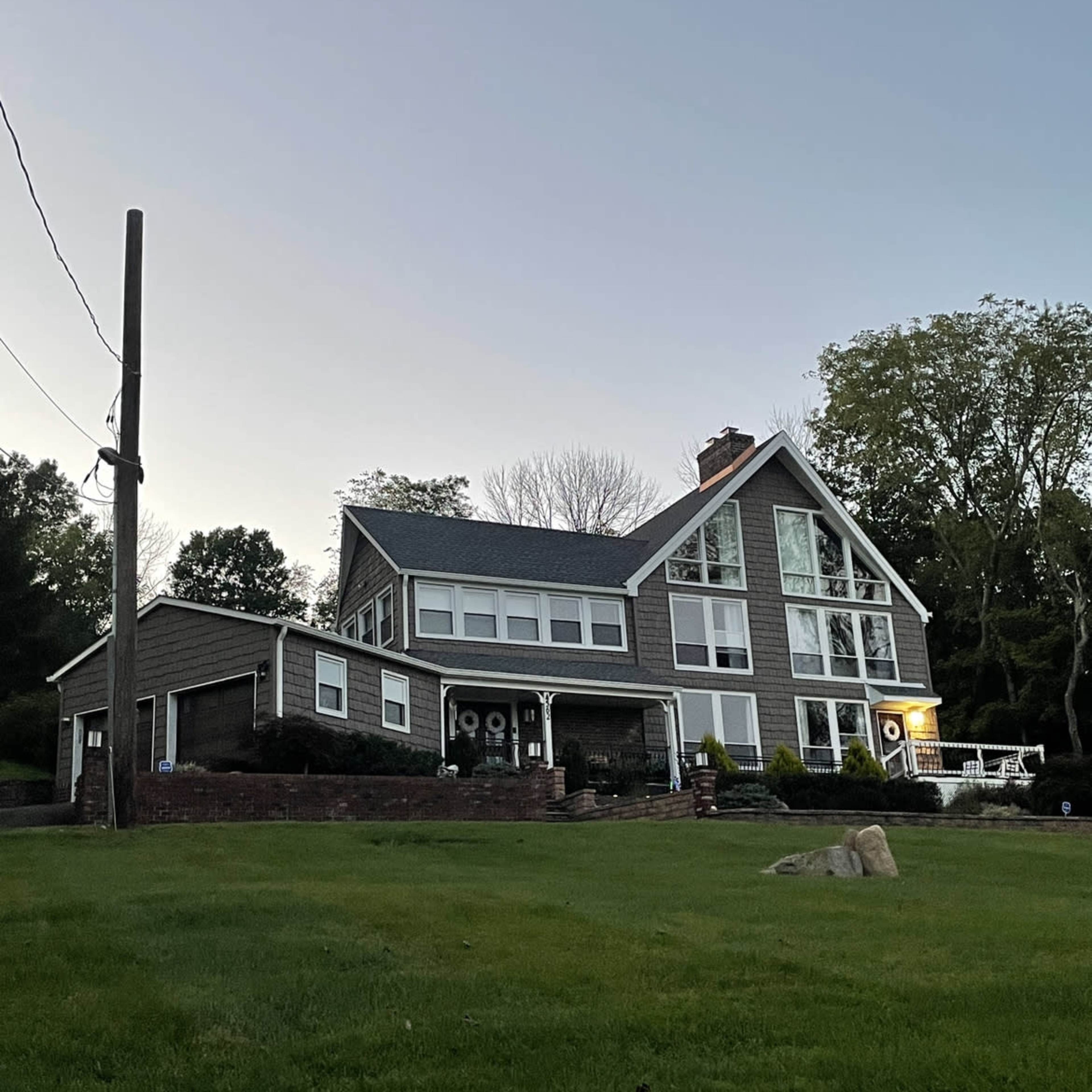 A large, modern house with multiple windows and a sloped roof is set on a grassy lawn bordered by a power pole and trees.