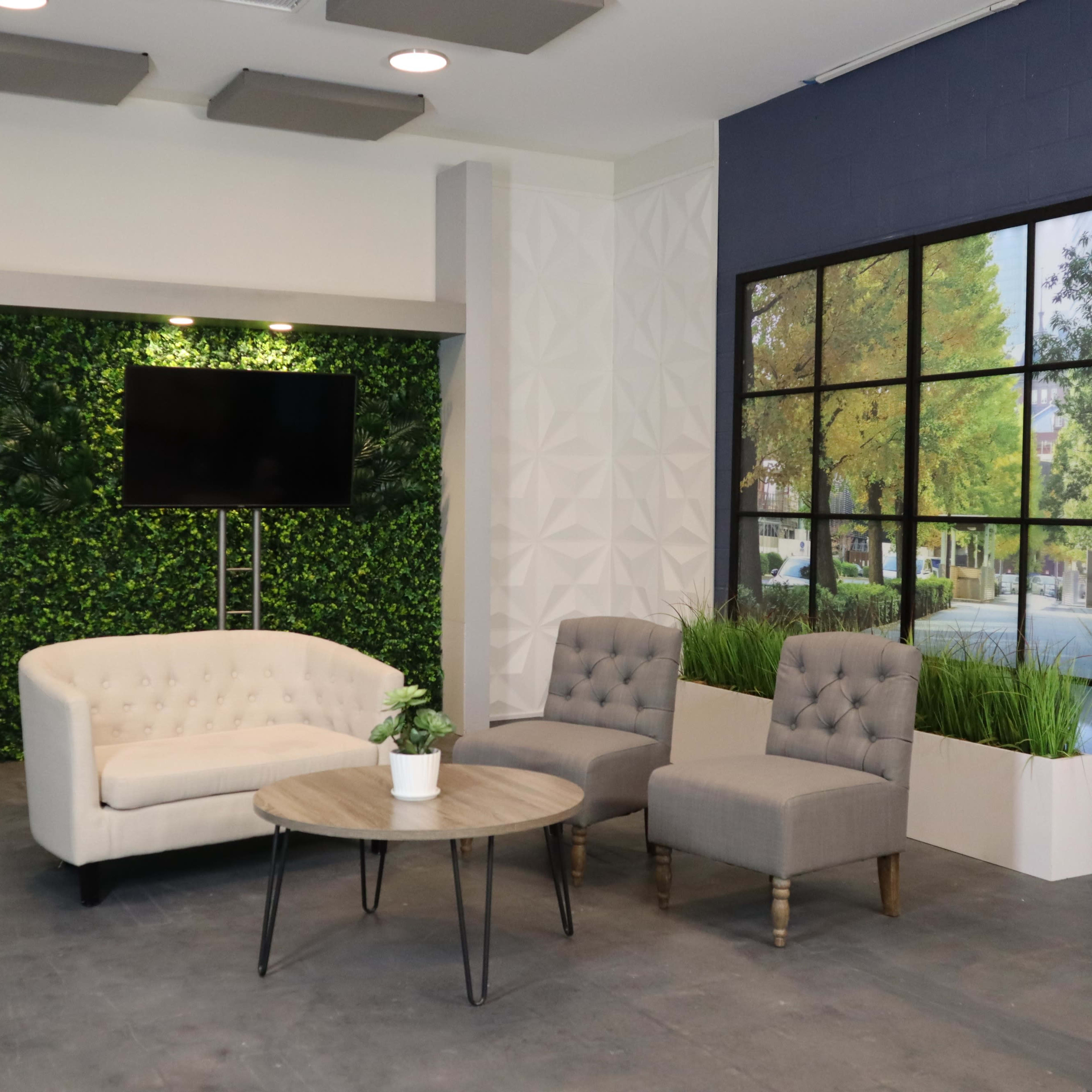 A modern lounge area with a white sofa, two gray chairs, a round wooden table, a television on a wall, and a large window graphic depicting a tree-lined street.
