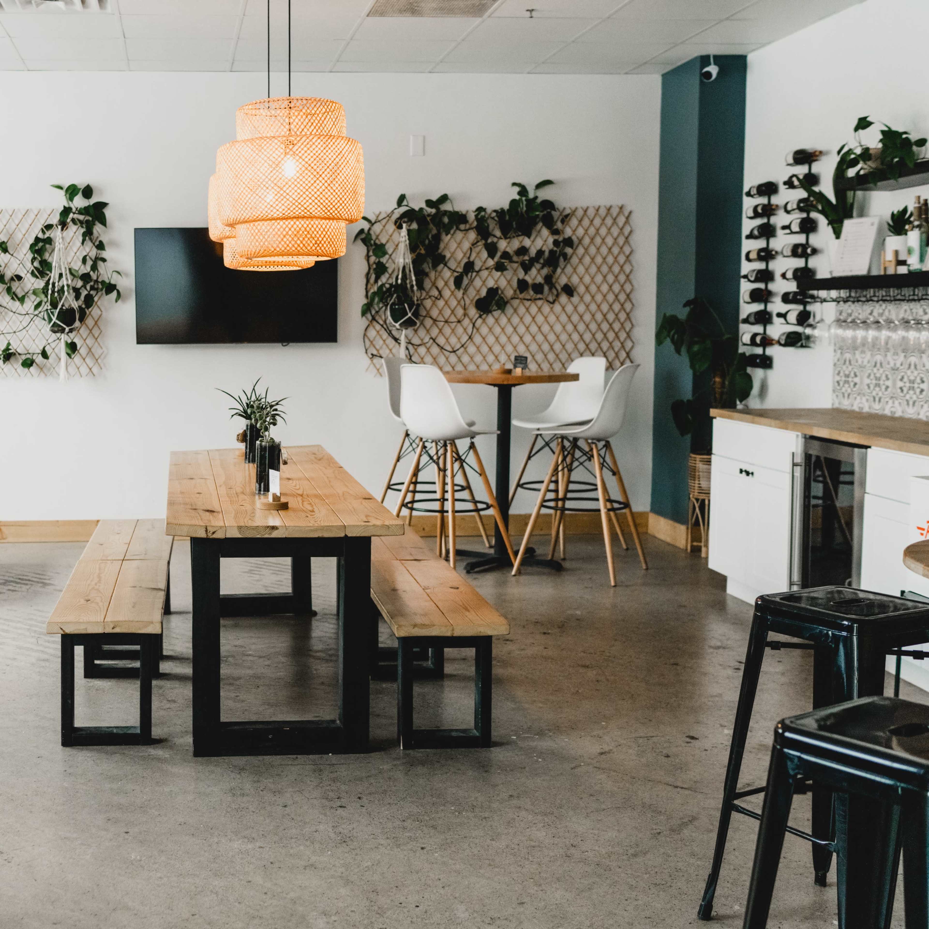 A modern café interior featuring wooden tables, a large pendant light, and greenery on the walls.