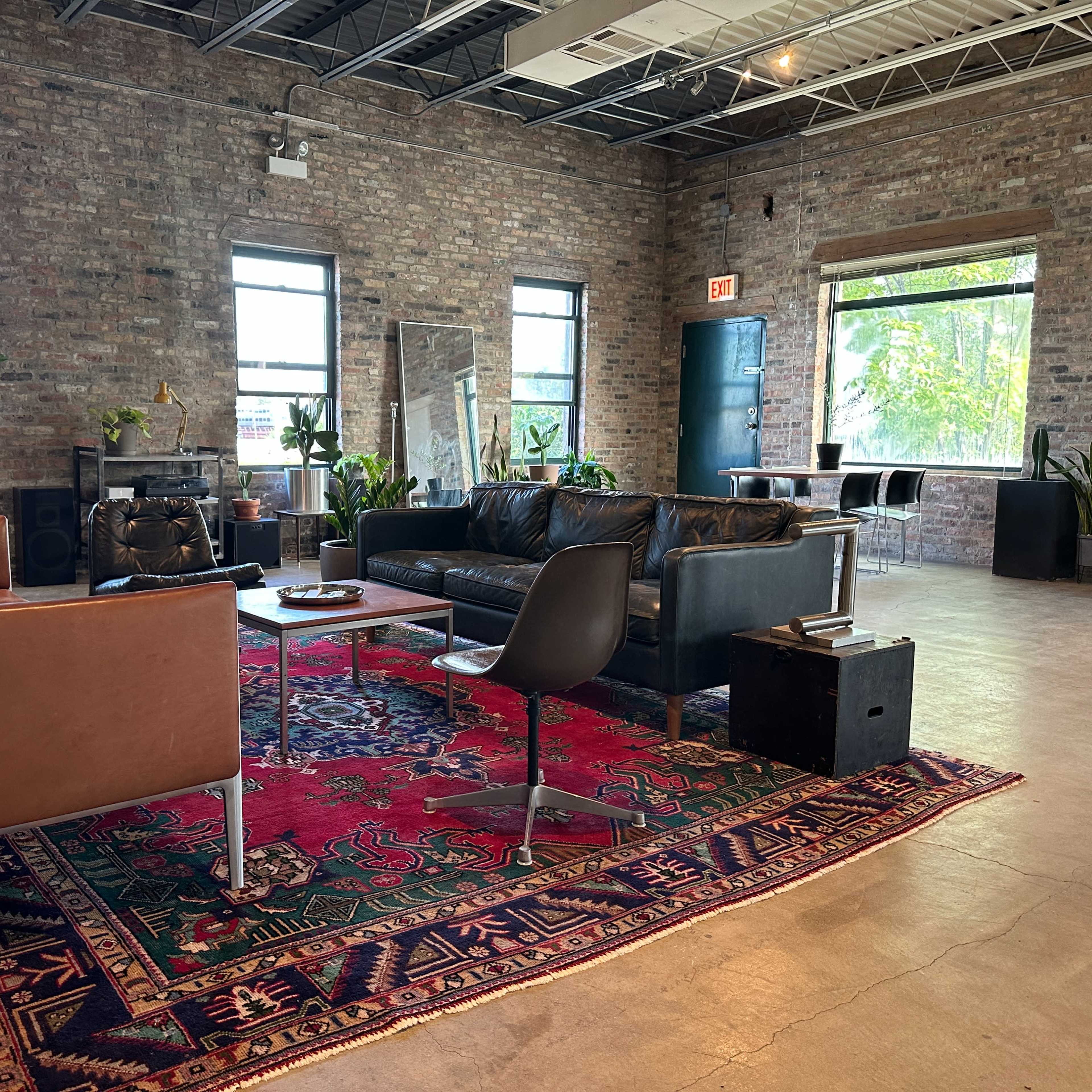 A spacious industrial-style living area with two sofas, a coffee table, and large windows allowing natural light, surrounded by plants and brick walls.