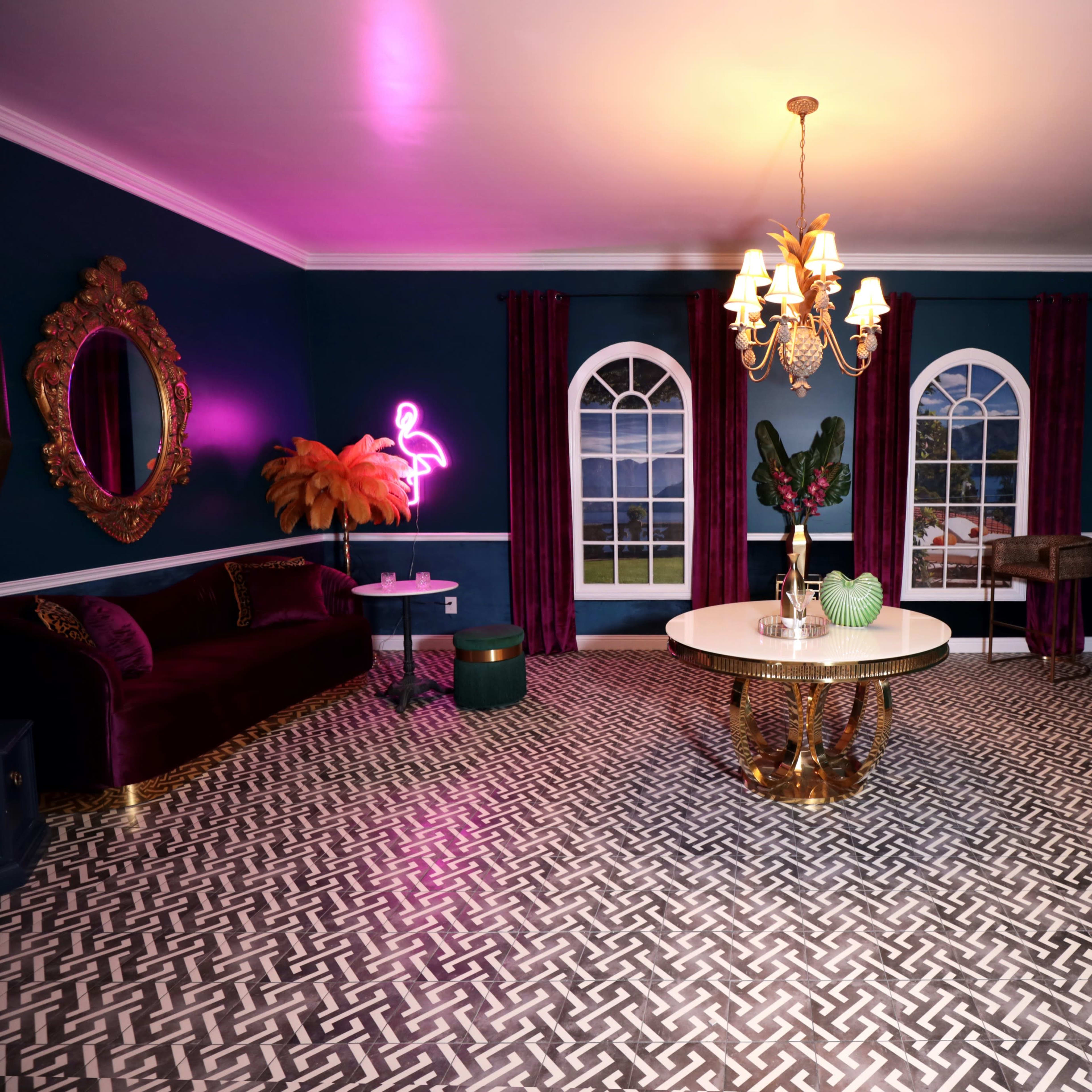 The interior of the room features a dark blue wall with a decorative chandelier, a round table with a gold base, a velvet couch, and large windows.