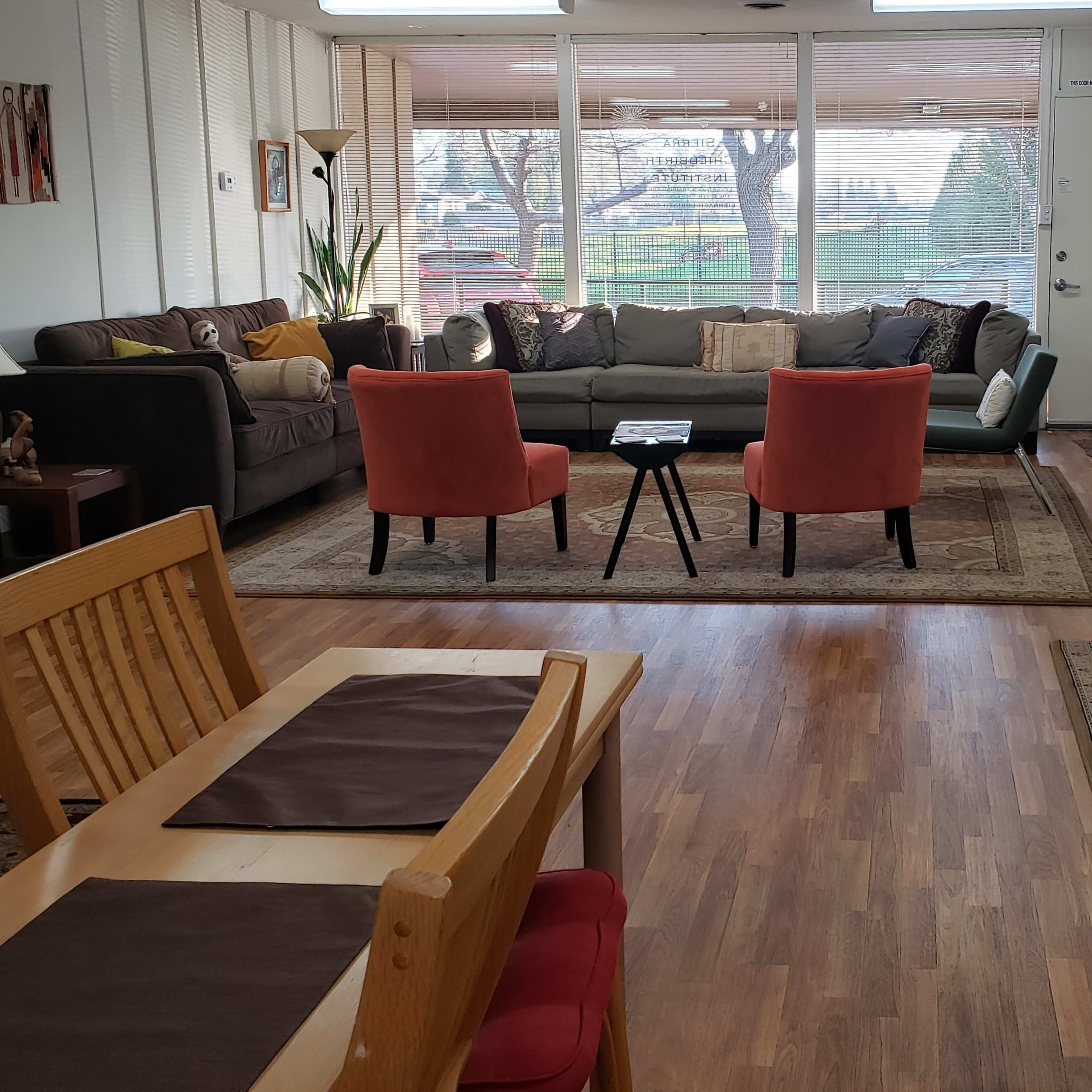 A cozy living space featuring a gray couch, two orange chairs, a wooden dining table, and a water dispenser against a backdrop of large windows.