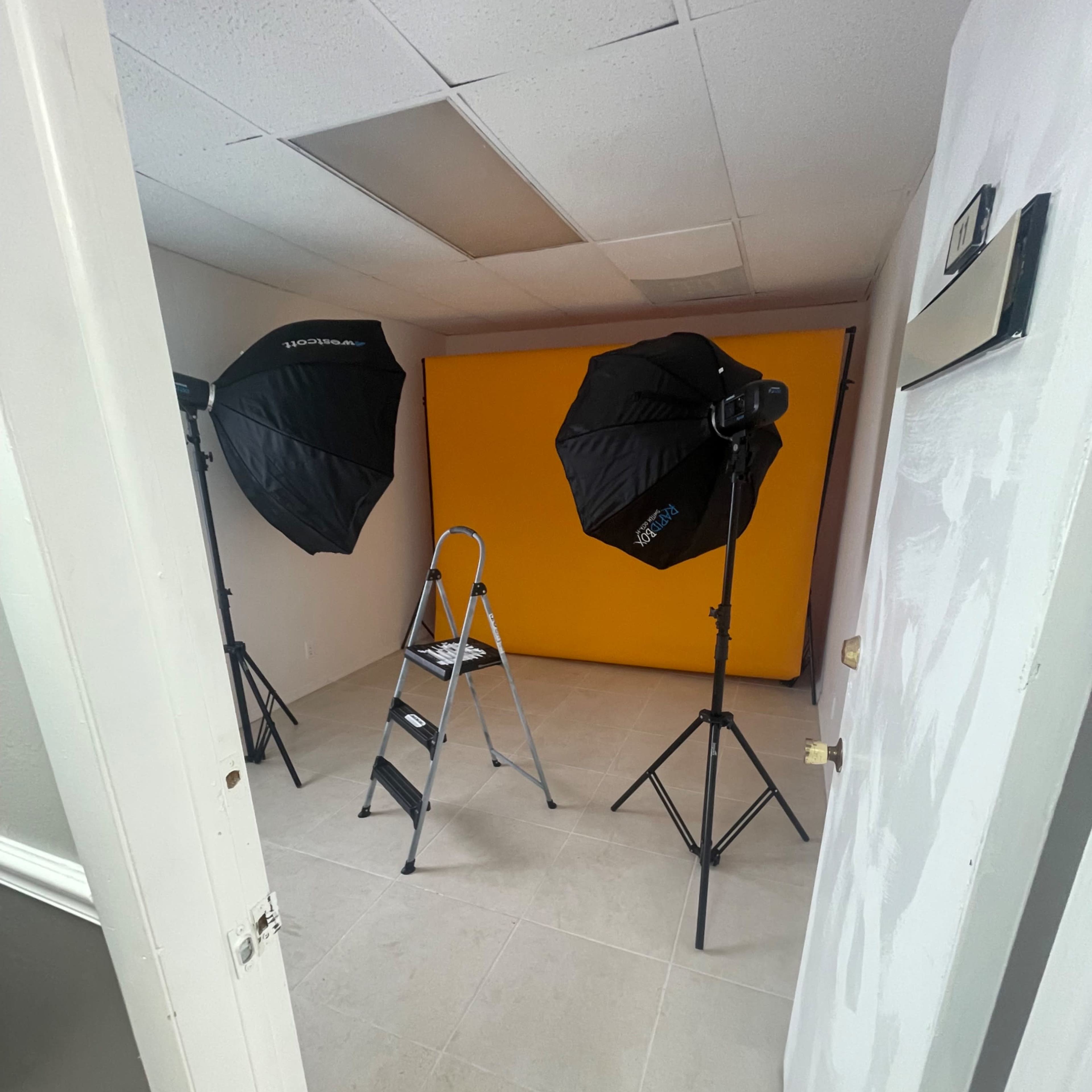 A photography studio setup with two umbrella lights, a step ladder, and a yellow backdrop.