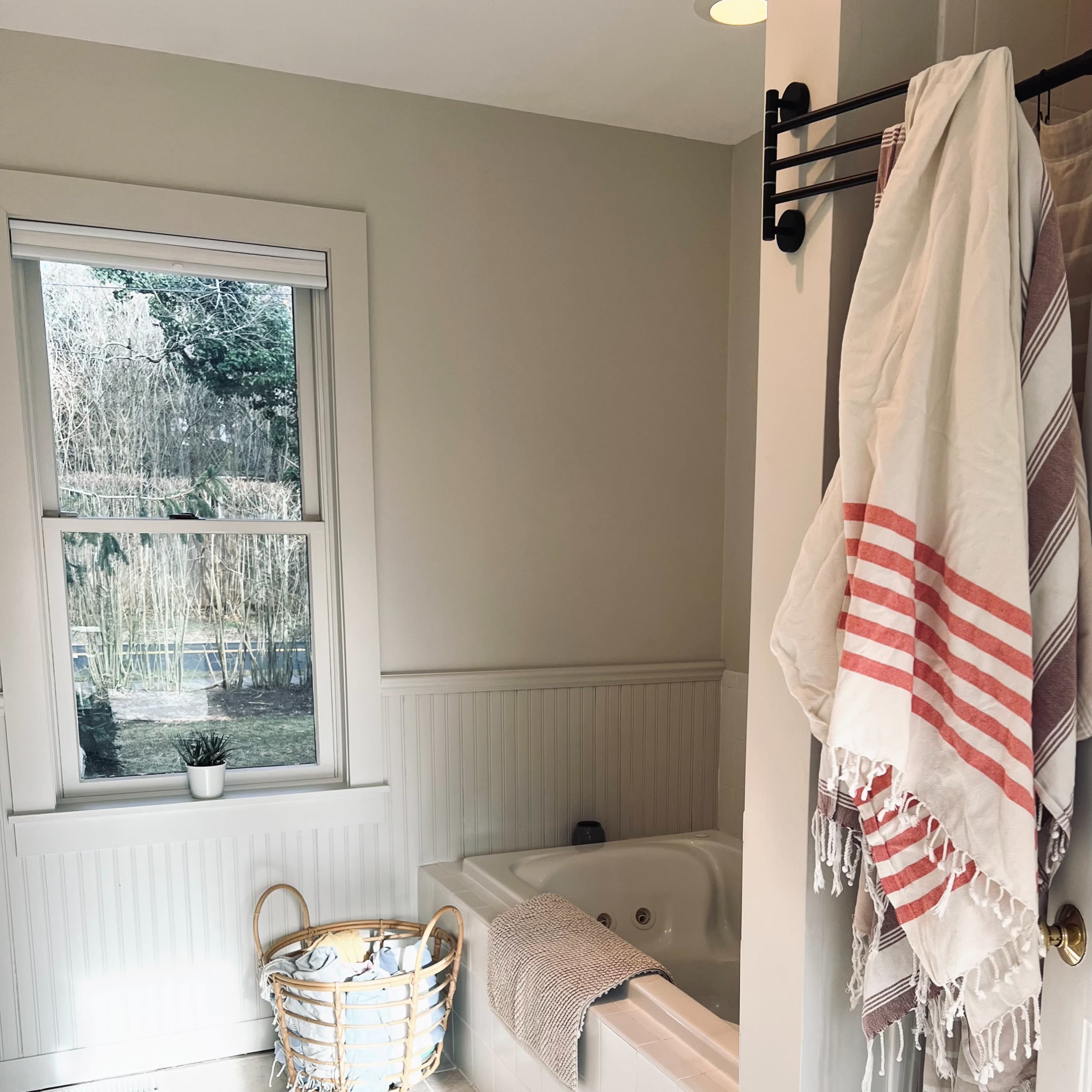 A bathroom with a large tub, a window overlooking greenery, a woven basket, and several towels on a rack.