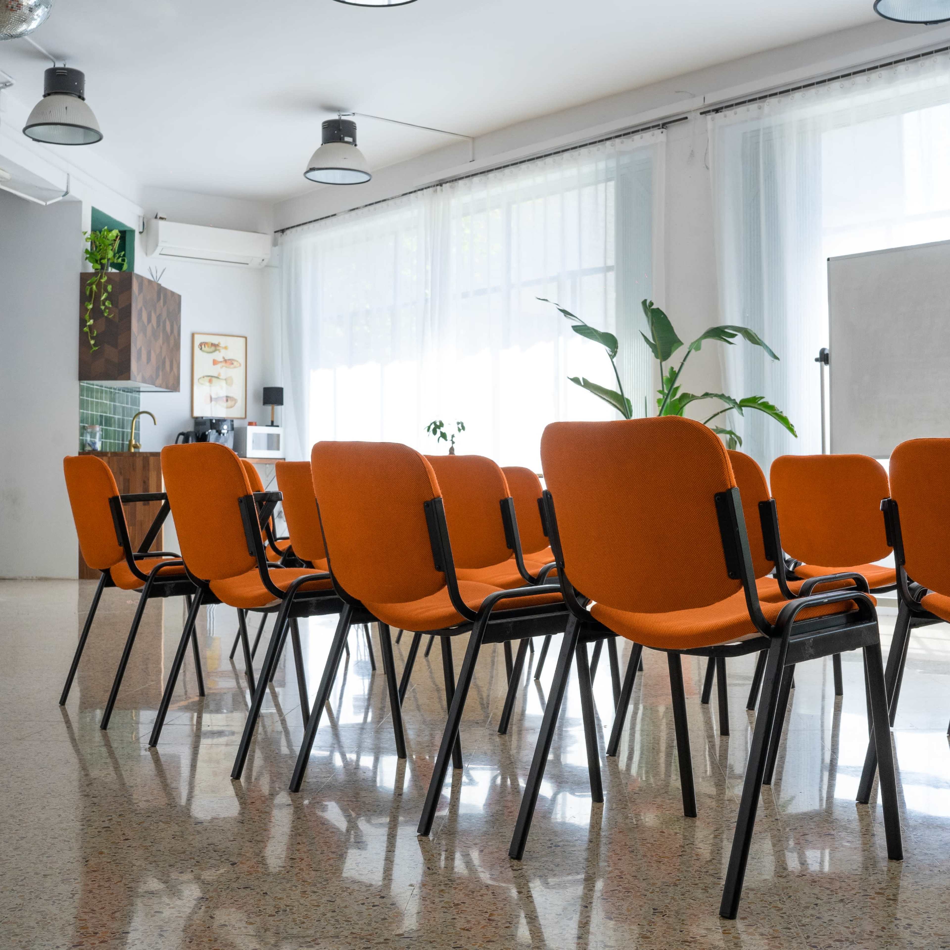 A row of orange chairs is arranged in a brightly lit room with large windows and a whiteboard in the background.