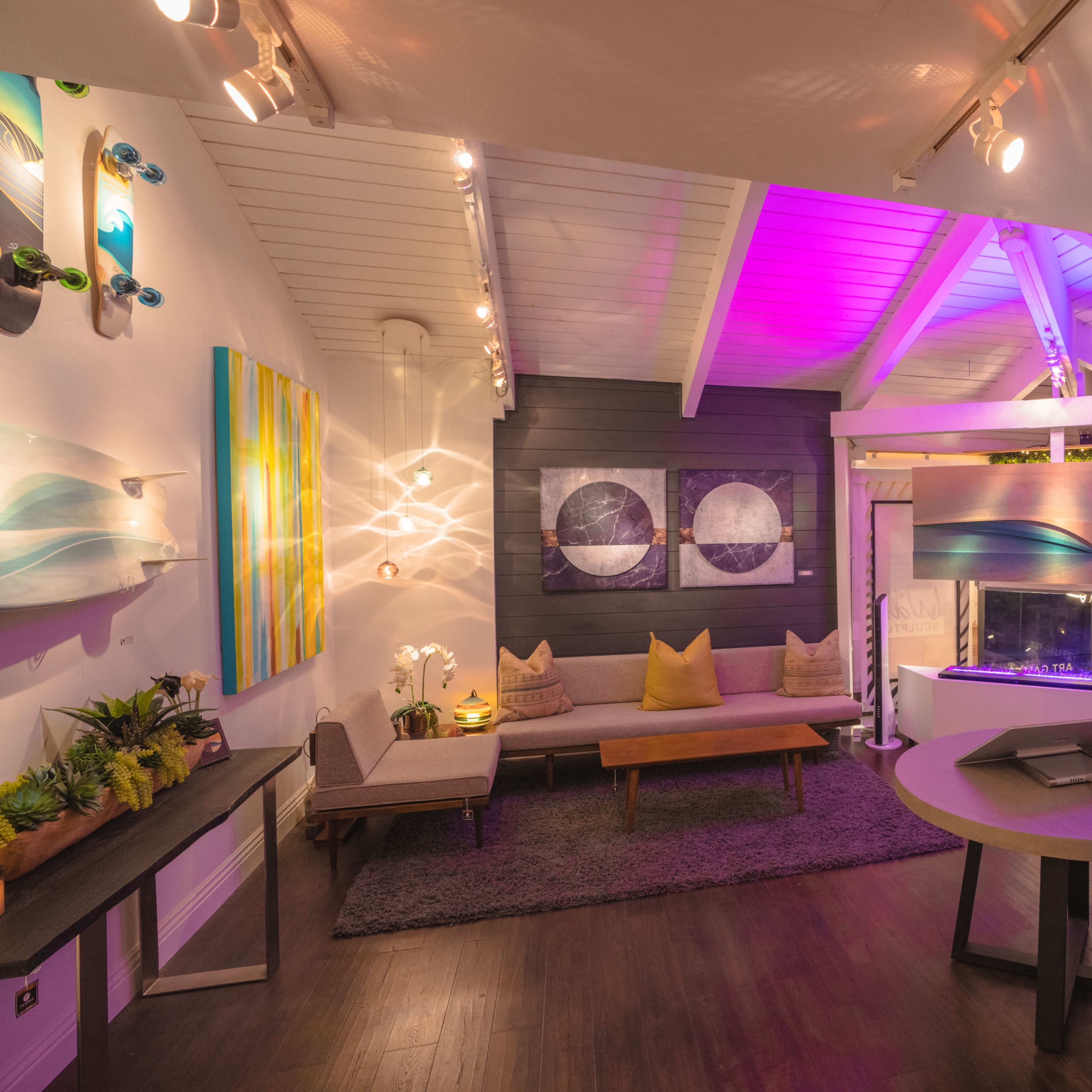 A modern, brightly lit art gallery with a mix of painted canvases and surfboard art displayed on the walls, along with a cozy seating area.