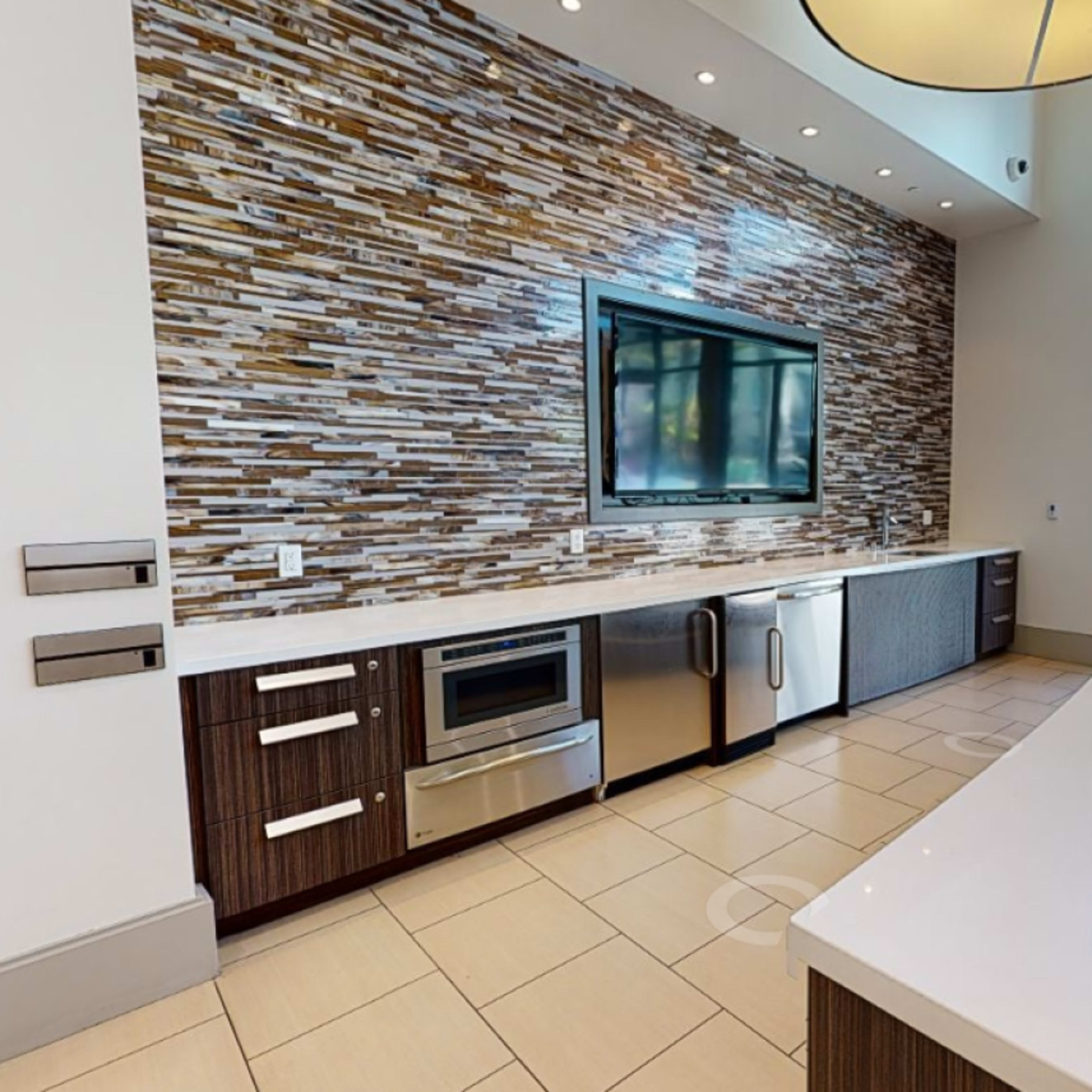 A modern kitchen area with a large countertop and a television mounted on the wall, adjacent to large windows overlooking a swimming pool.