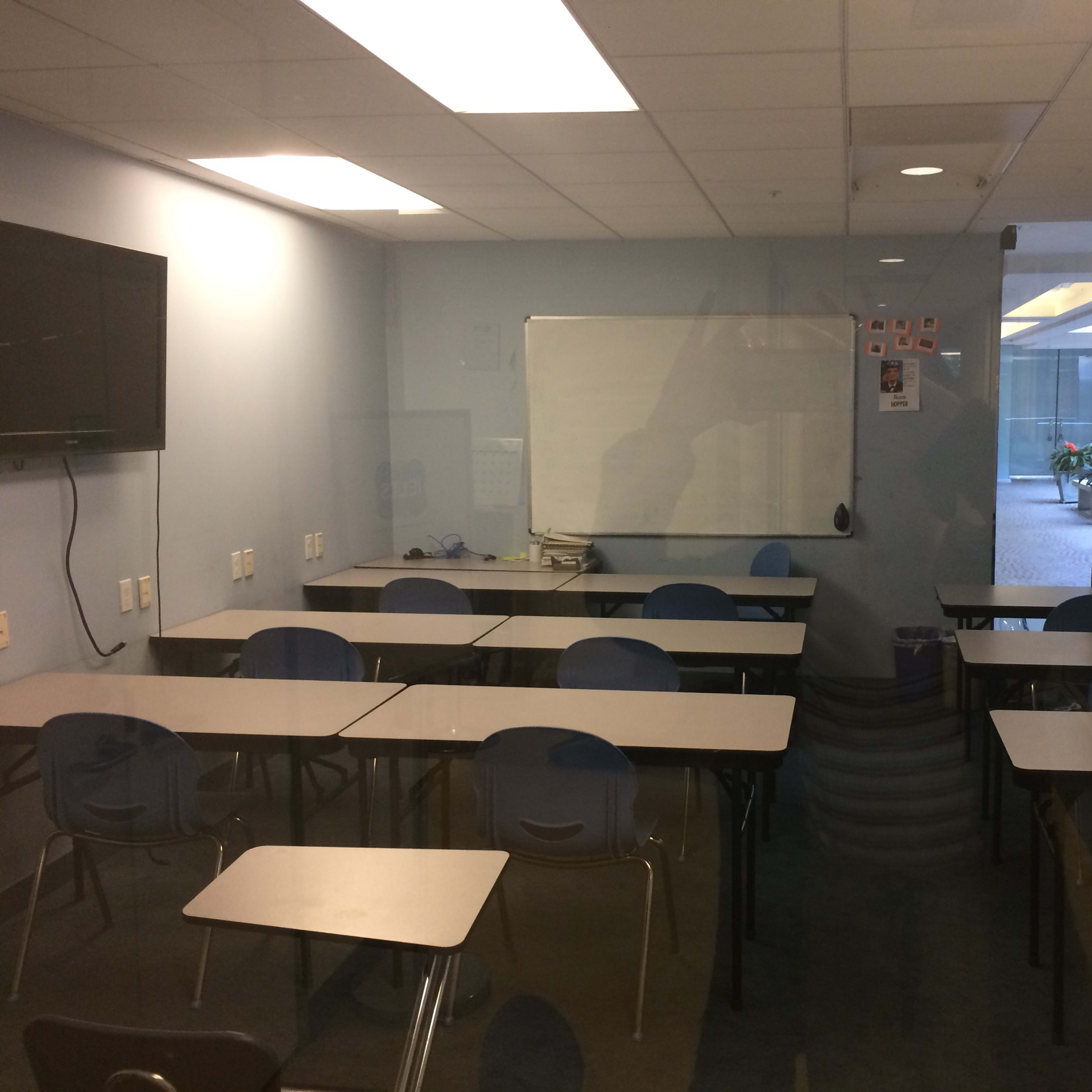 A classroom is depicted with rows of empty desks facing a whiteboard and a television mounted on the wall.