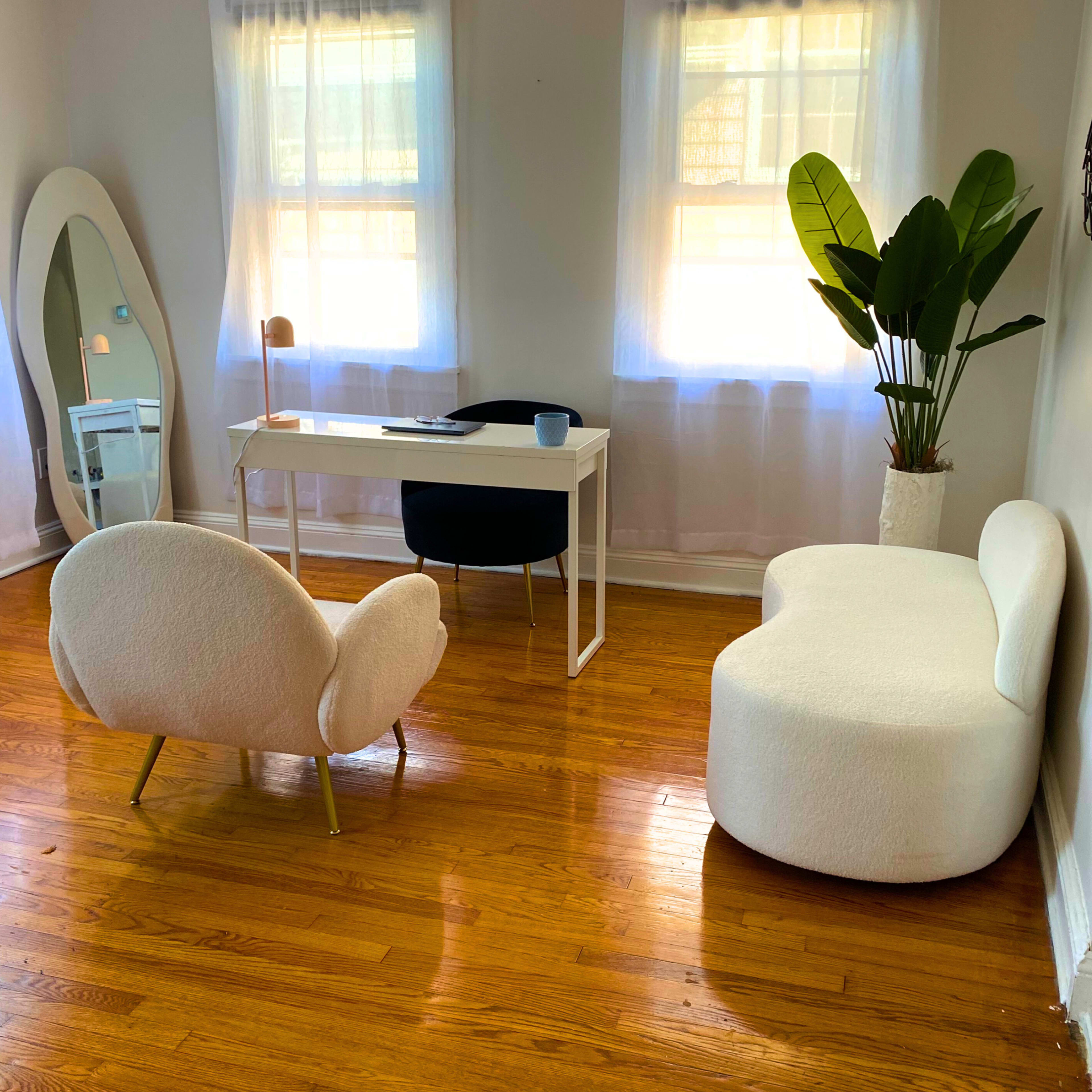 A bright room featuring a white desk with a chair, two modern chairs, a large mirror, and a potted plant.