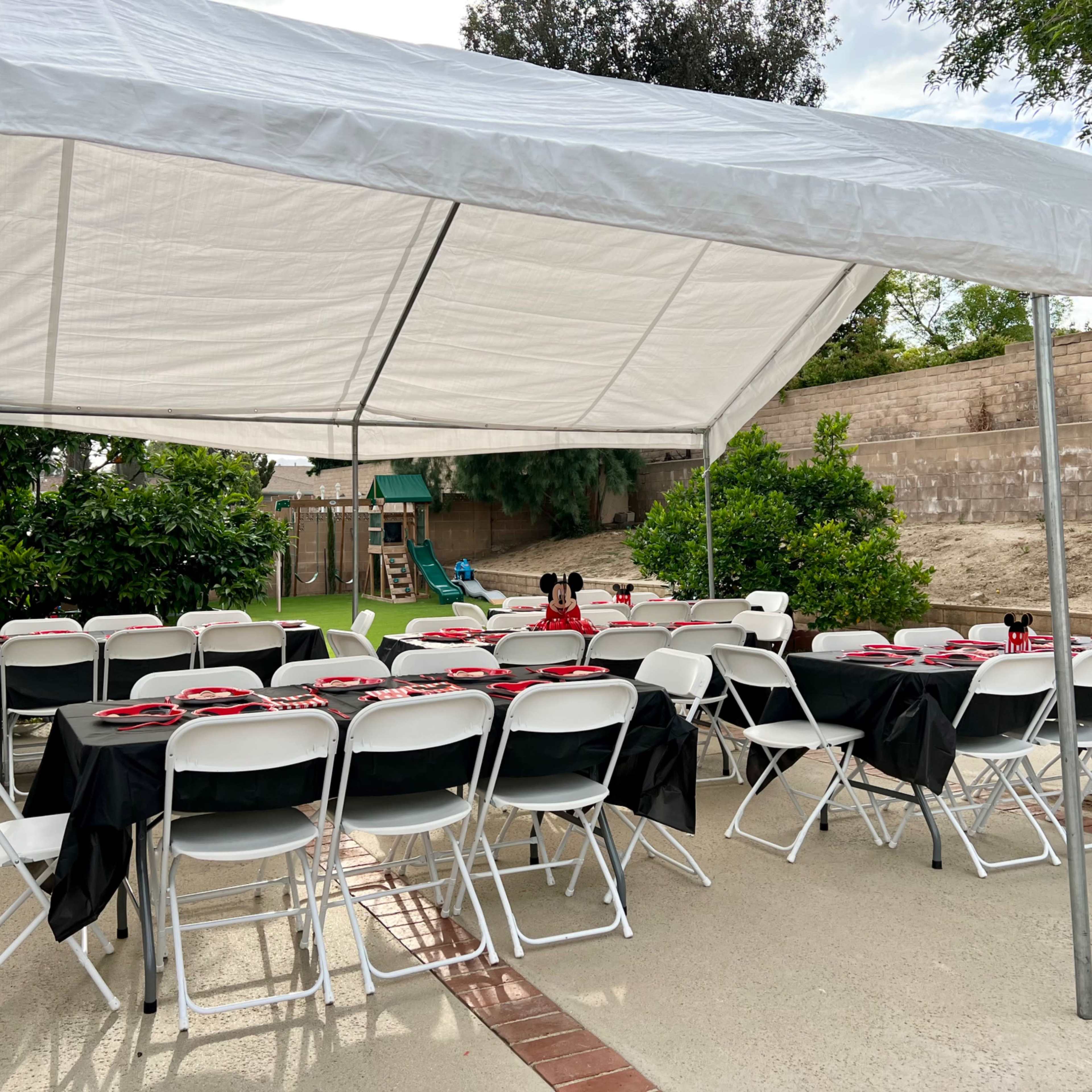 A large tent is set up in a backyard, containing tables covered with black cloths and red plates, while a playground structure is visible in the background.