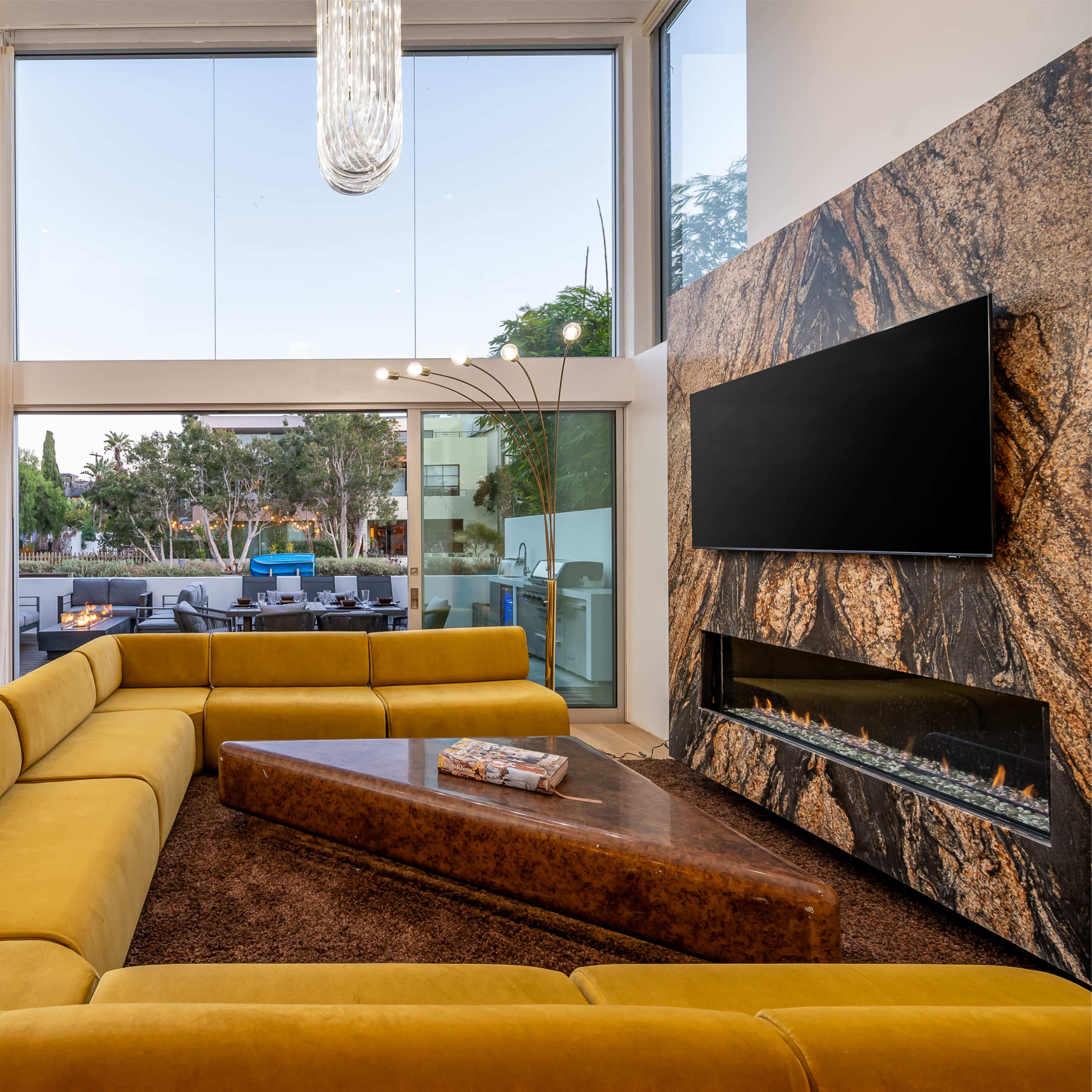 The interior features a spacious living room with a large yellow sectional sofa, a modern fireplace, and floor-to-ceiling windows providing a view of an outdoor patio and greenery.