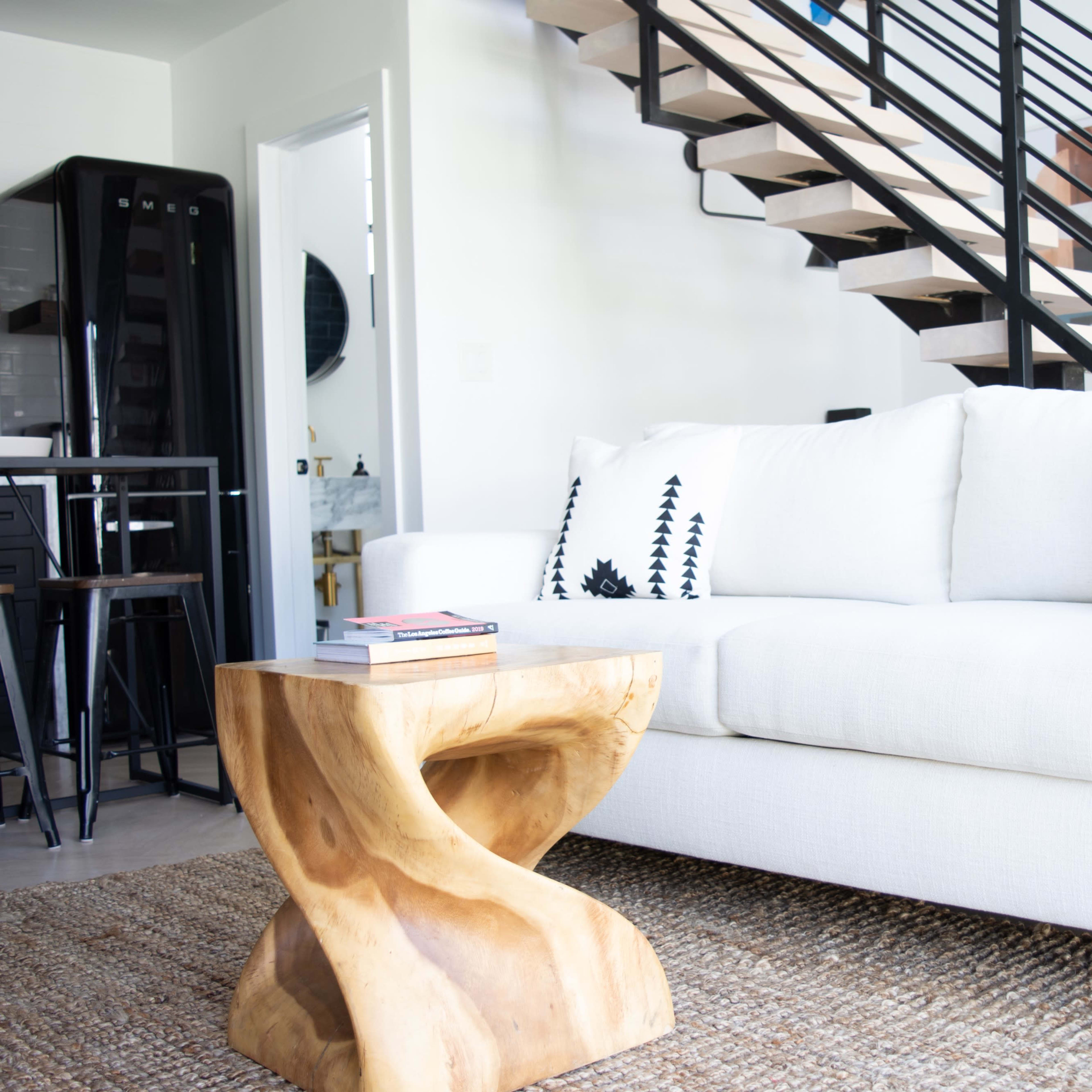 A modern living space featuring a white sofa with patterned throw pillows, a wooden sculpture table, and a staircase with a black railing in the background.