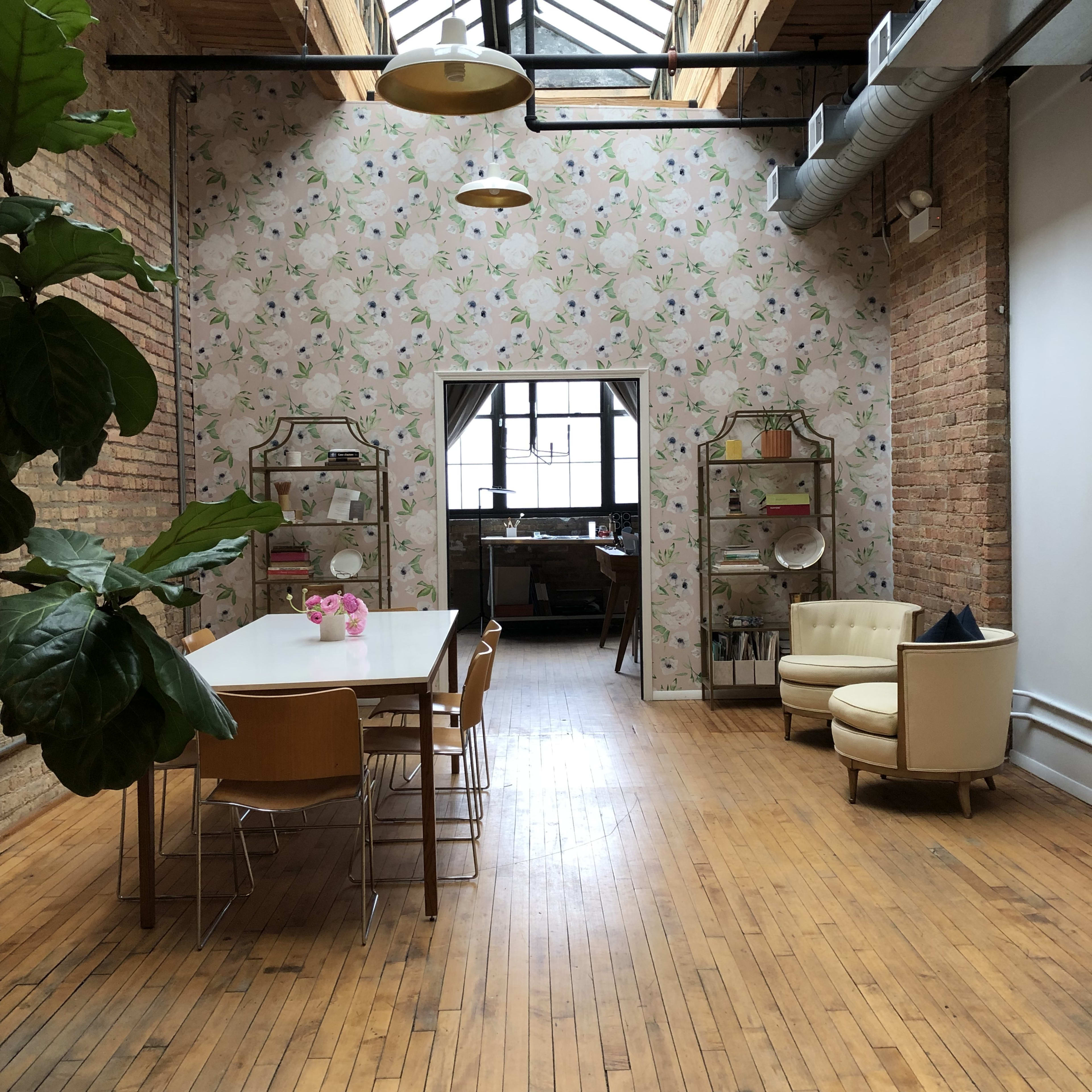 A spacious, well-lit room featuring a long dining table surrounded by chairs, a cozy seating area with an armchair, and a decorative shelf against a floral wallpaper backdrop.