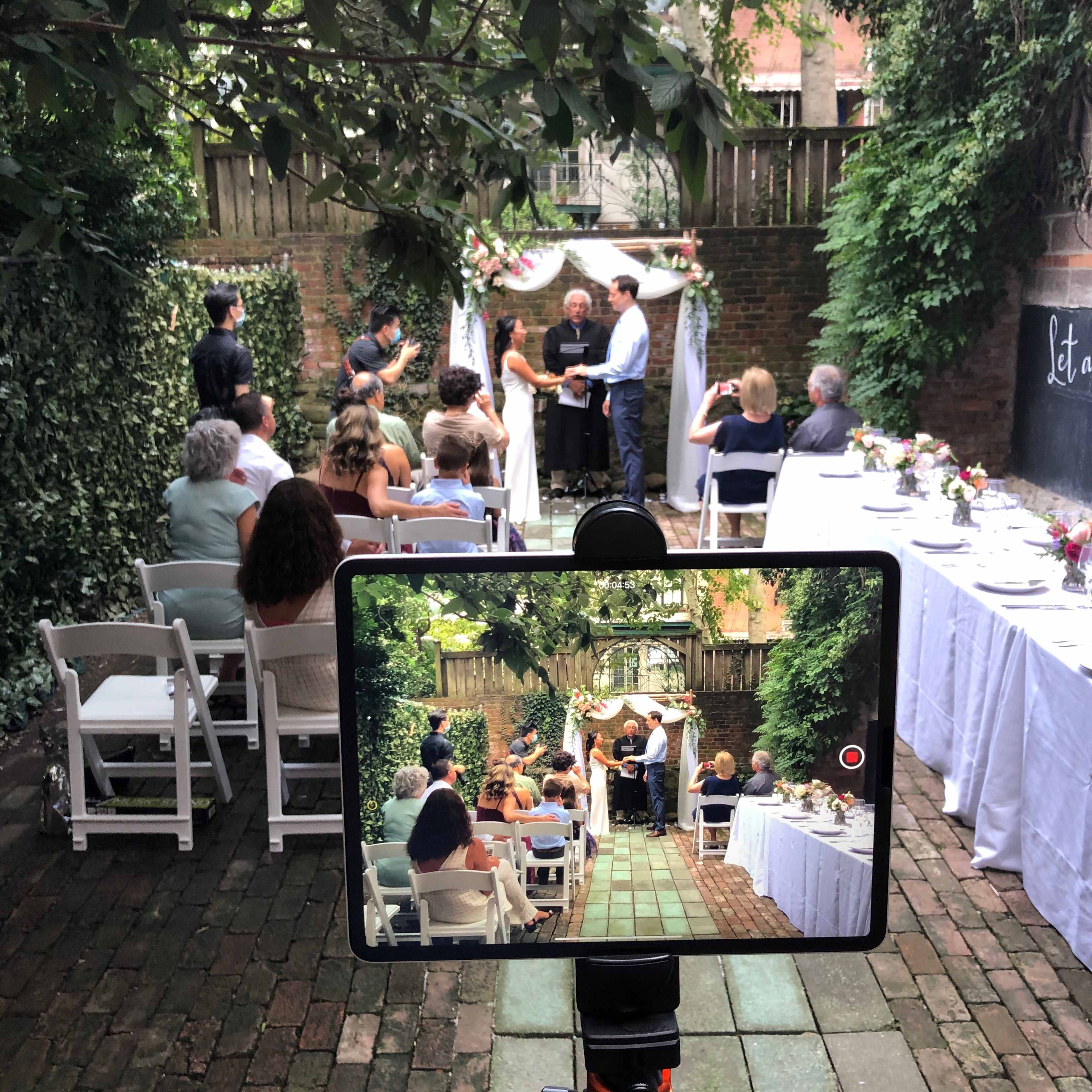 A couple stands under a floral arch during a wedding ceremony in a garden, while guests are seated at tables nearby.