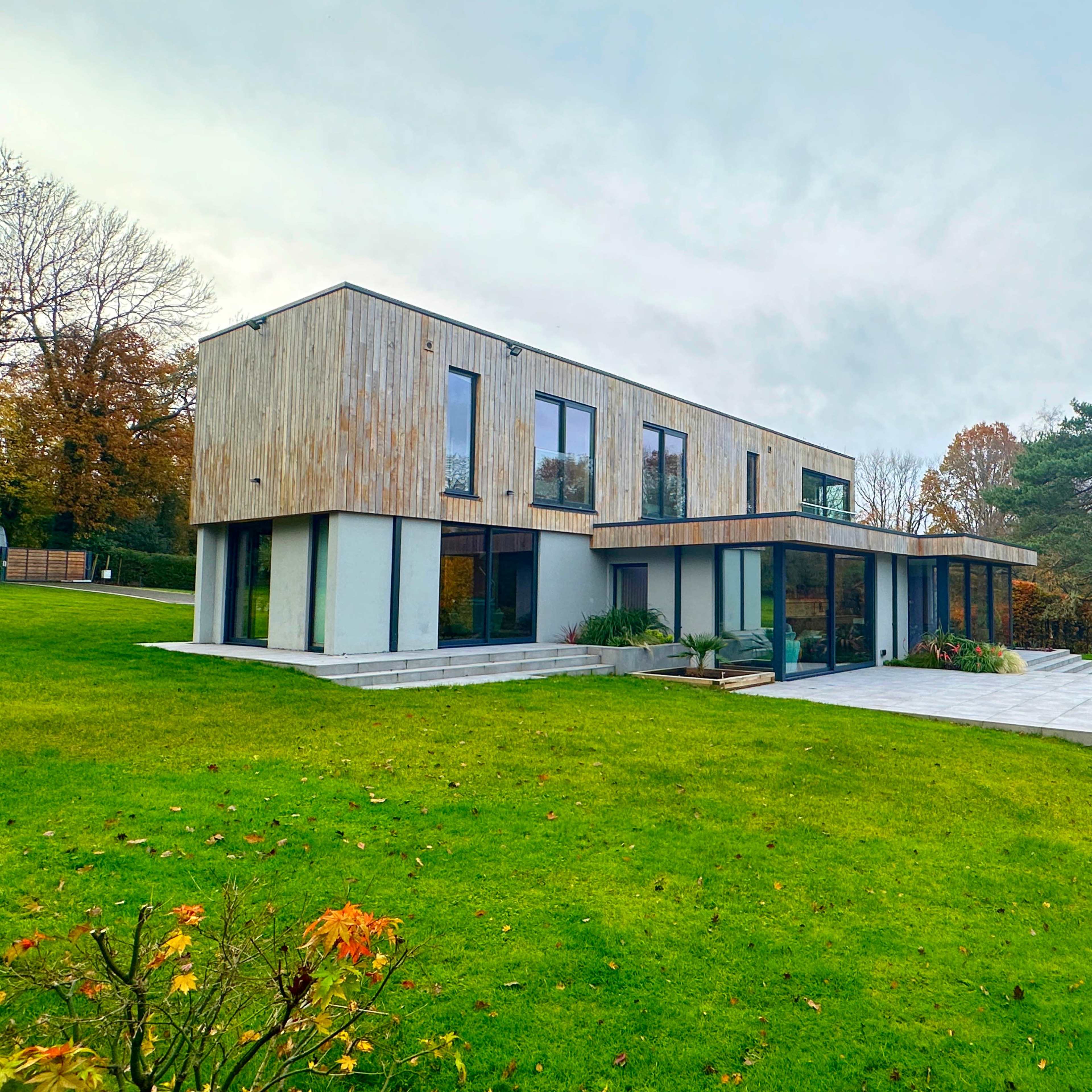 A modern two-story house with a wooden facade and large windows is situated on a green lawn surrounded by trees.