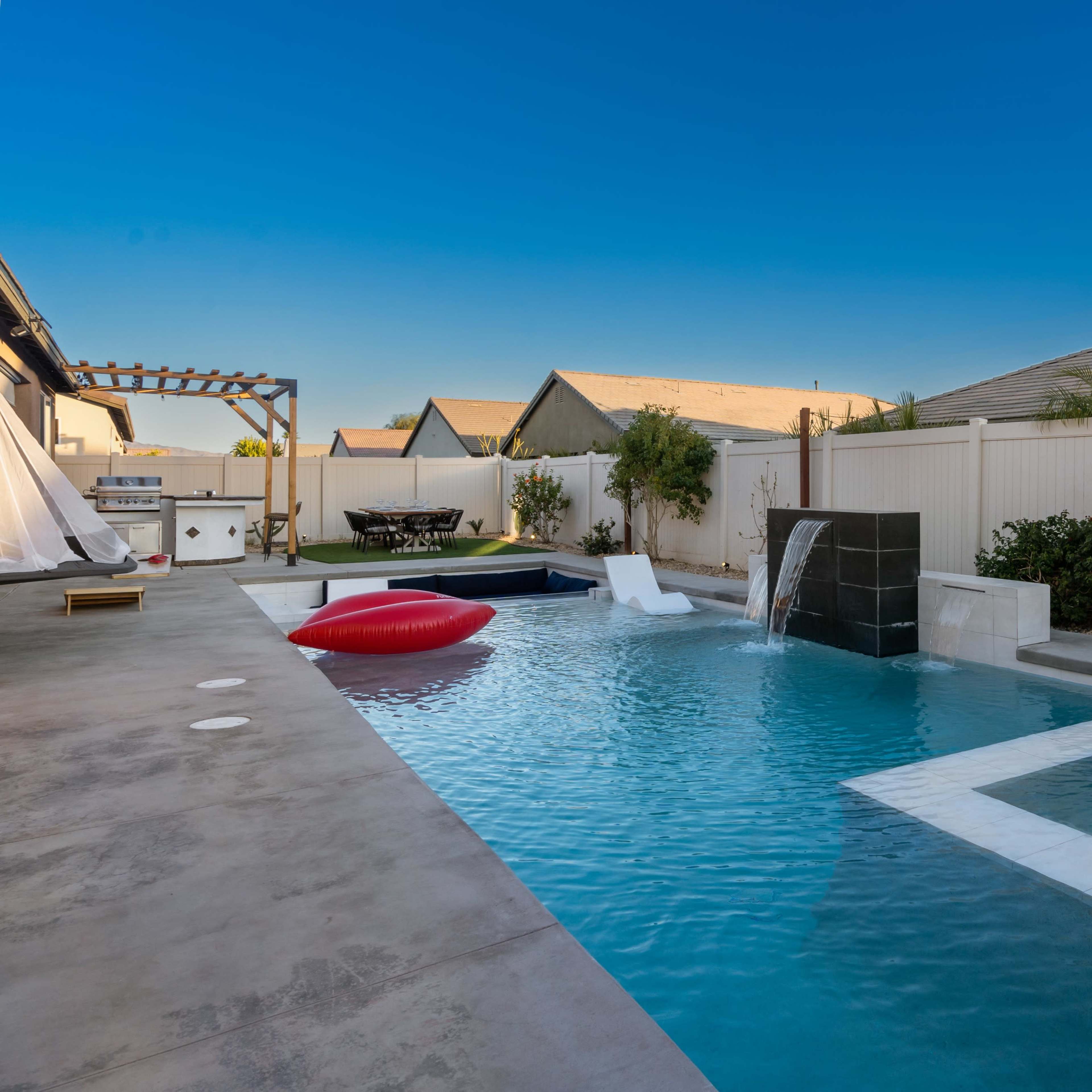 A modern backyard with a swimming pool, a wall fountain, and a seating area surrounded by houses and landscaping.