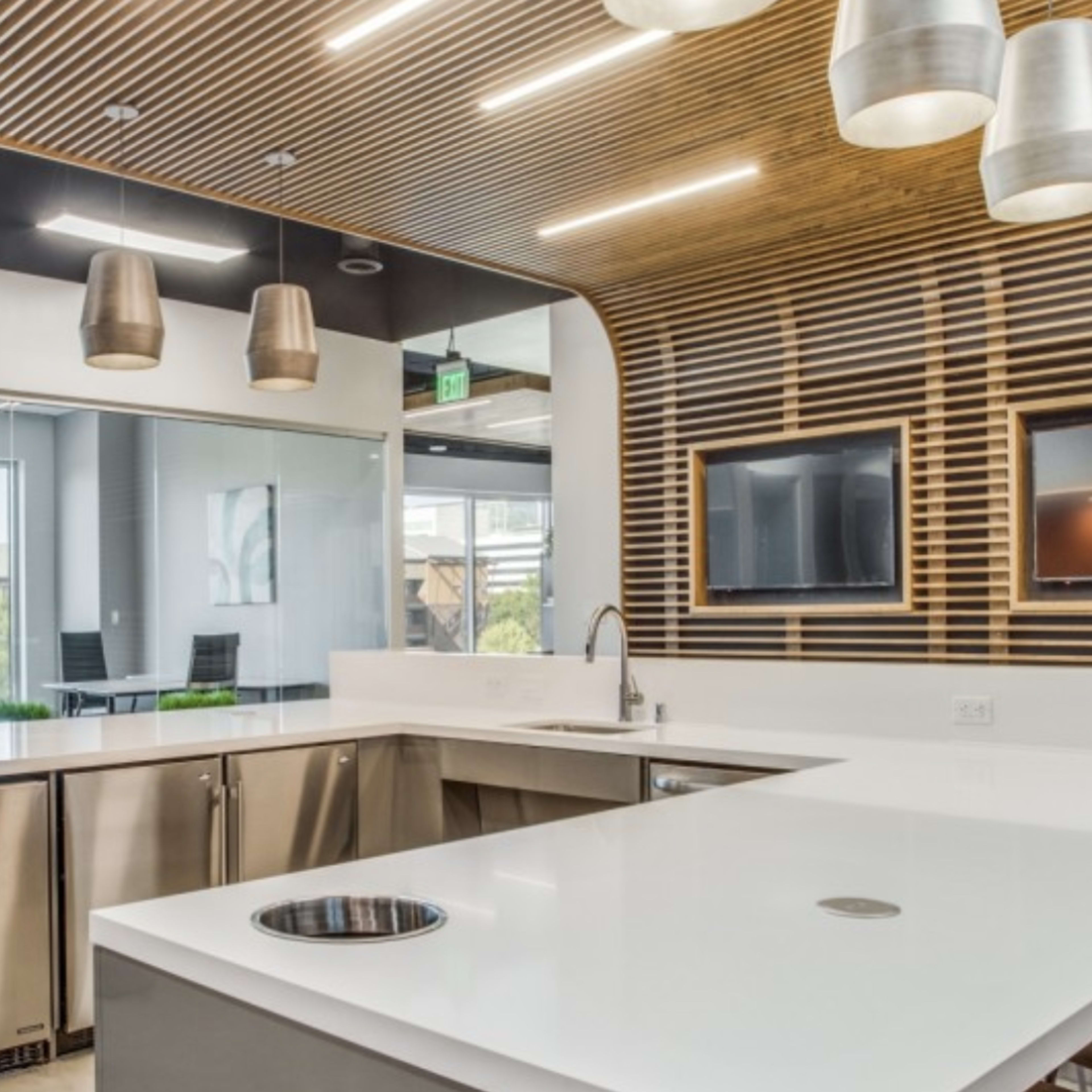 A modern kitchen area with stainless steel appliances, a white countertop, and large pendant lights, adjacent to a meeting space with large windows.