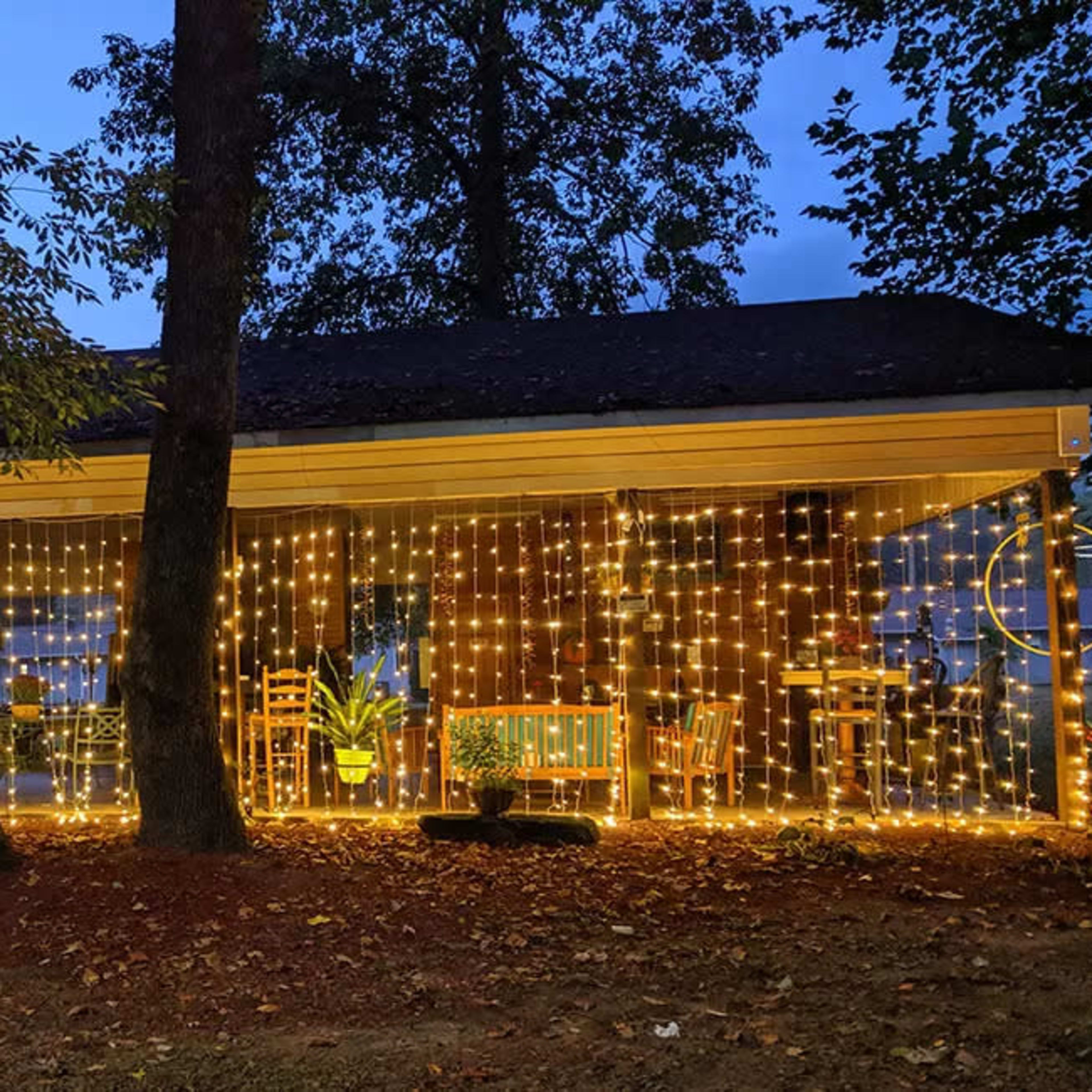 A porch illuminated by decorative string lights, surrounded by trees and a few chairs.