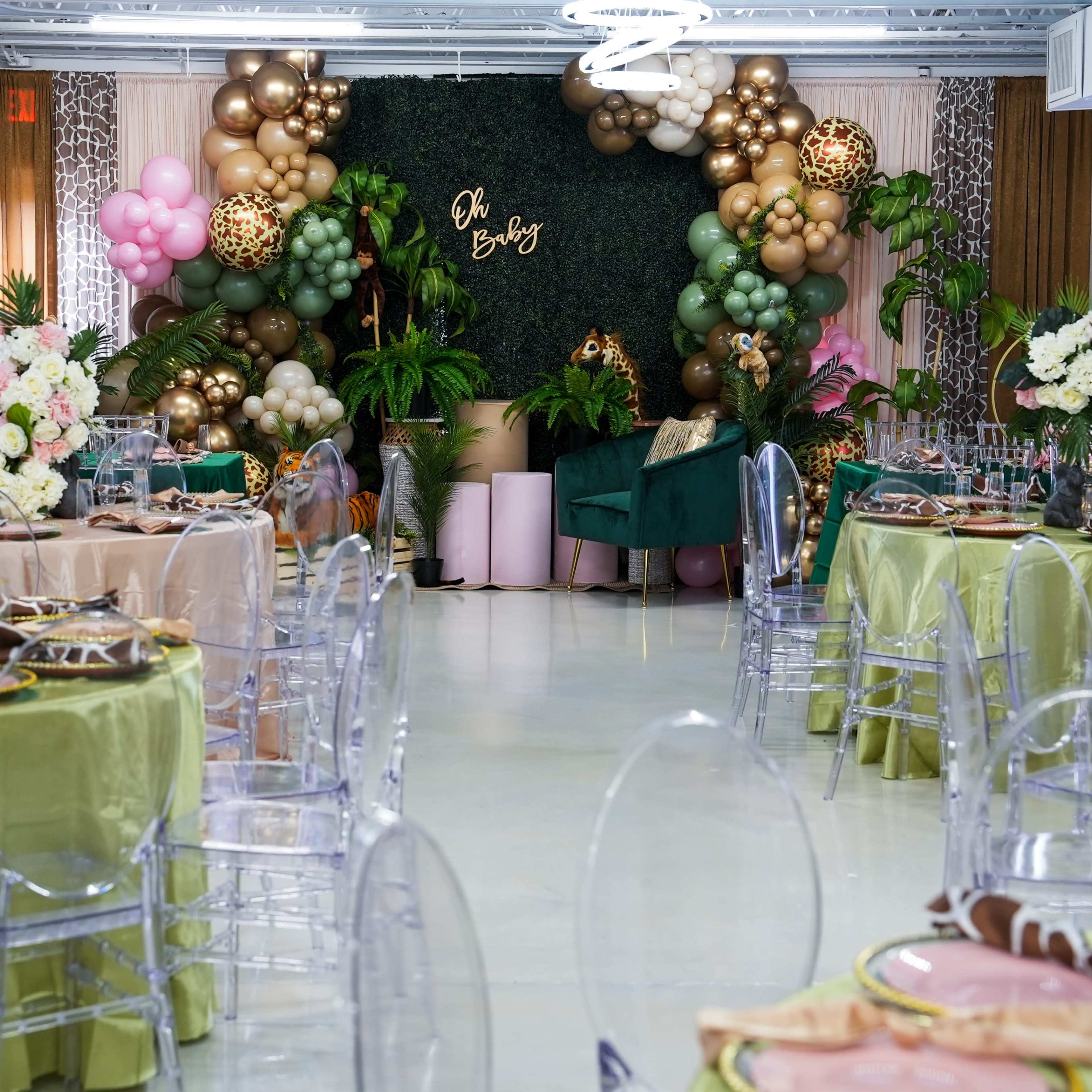 An elegantly decorated event space featuring clear chairs, round tables with various colored tablecloths, and a backdrop of balloons and greenery.