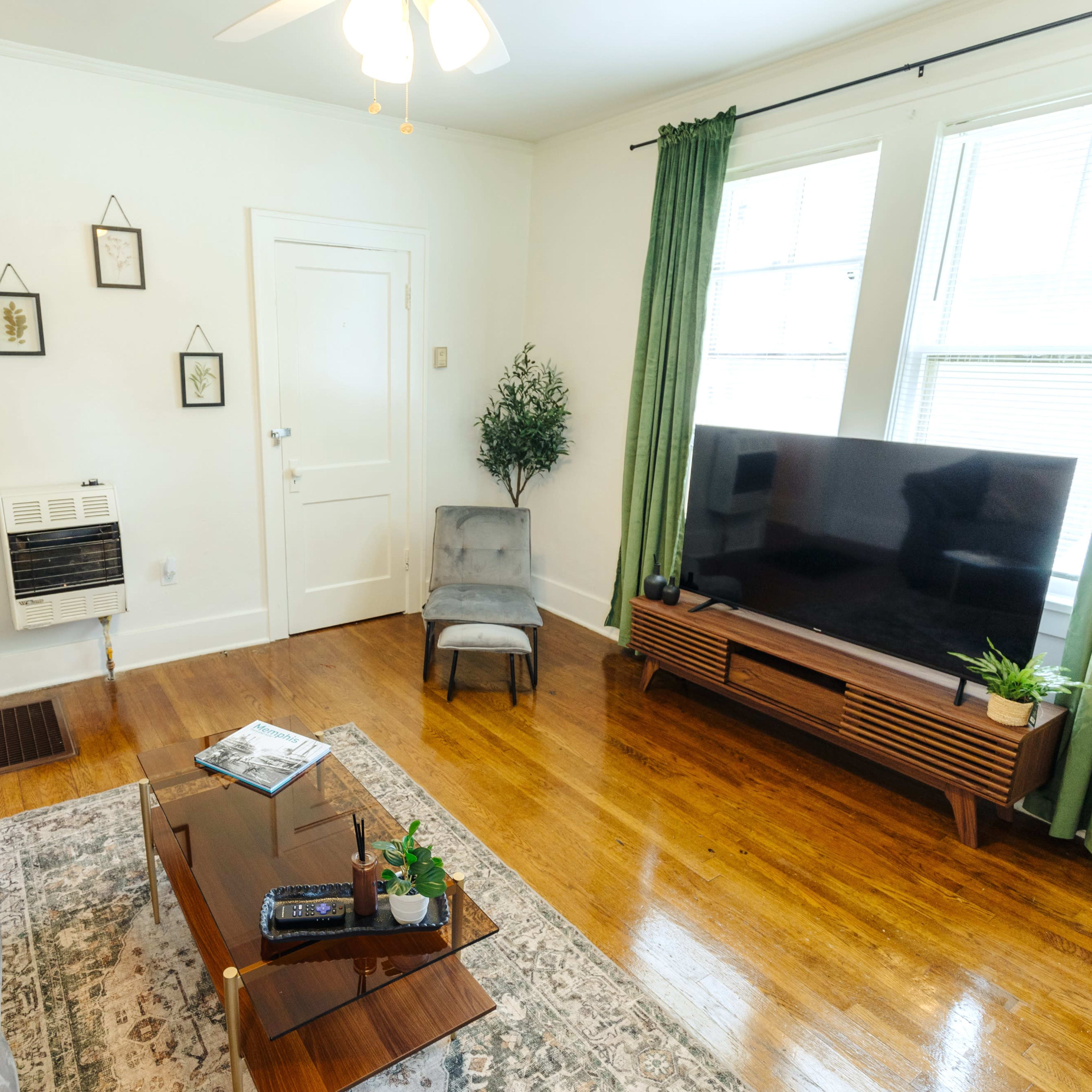 A living room with a sofa, a coffee table, a television stand, and large windows dressed with green curtains.