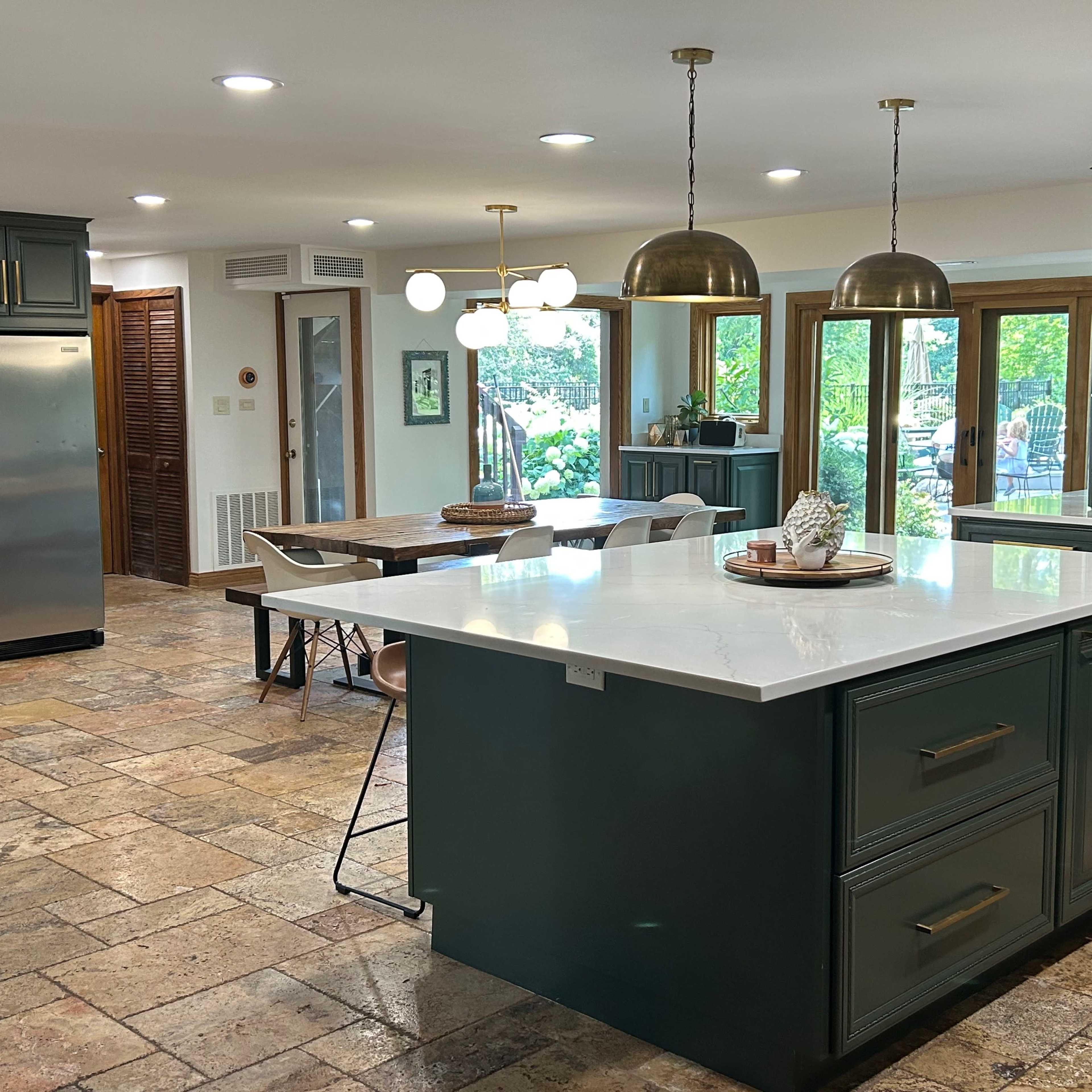 A spacious kitchen with an island, dark green cabinetry, stone flooring, and large windows allowing natural light.