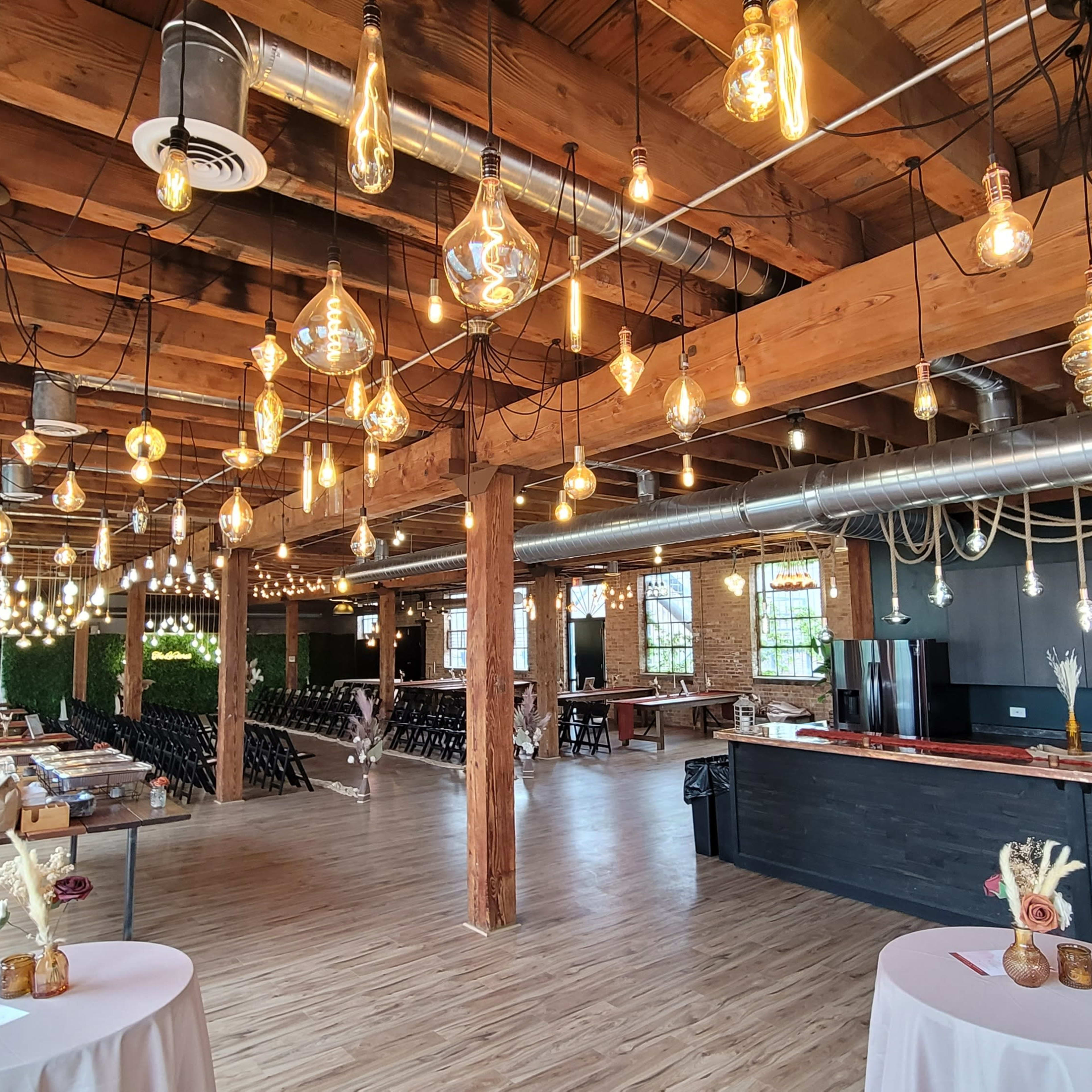 A spacious banquet hall with wooden beams, hanging Edison bulbs, and a bar area along one side, set up for an event.