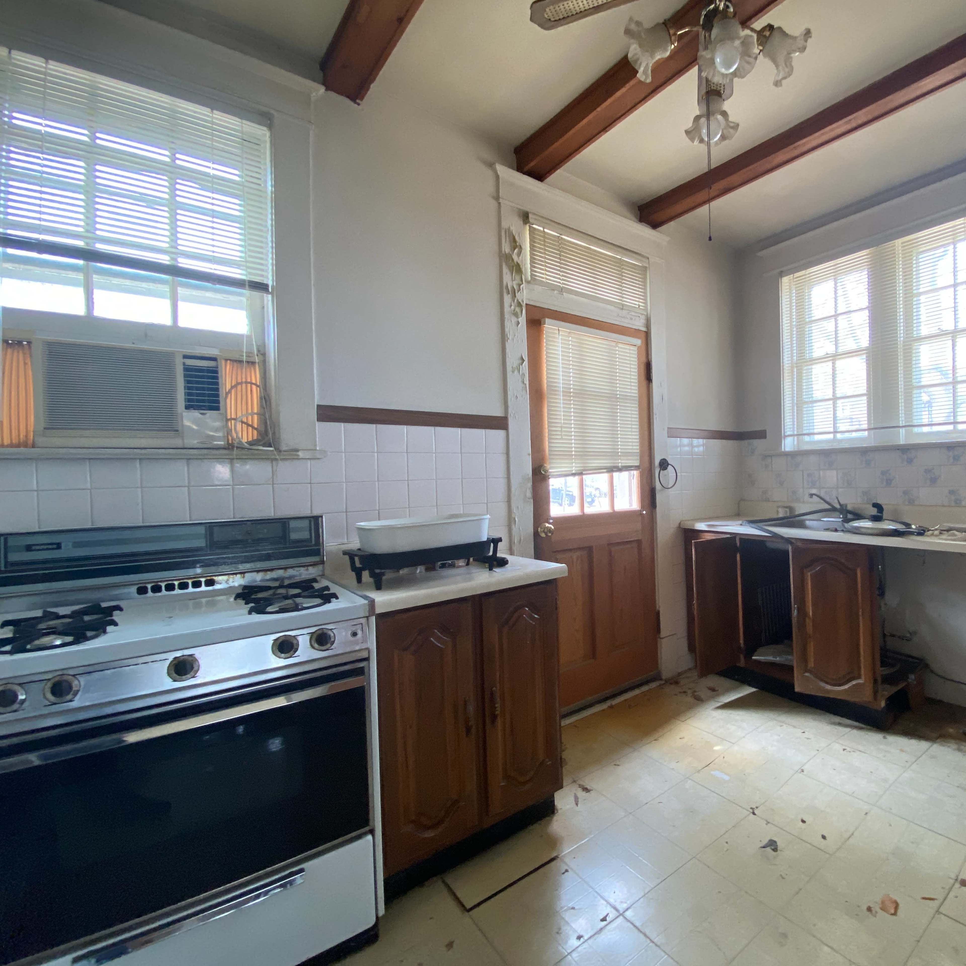 An empty kitchen with a stove, wooden cabinets, and a door leading outside, illuminated by natural light from a window.