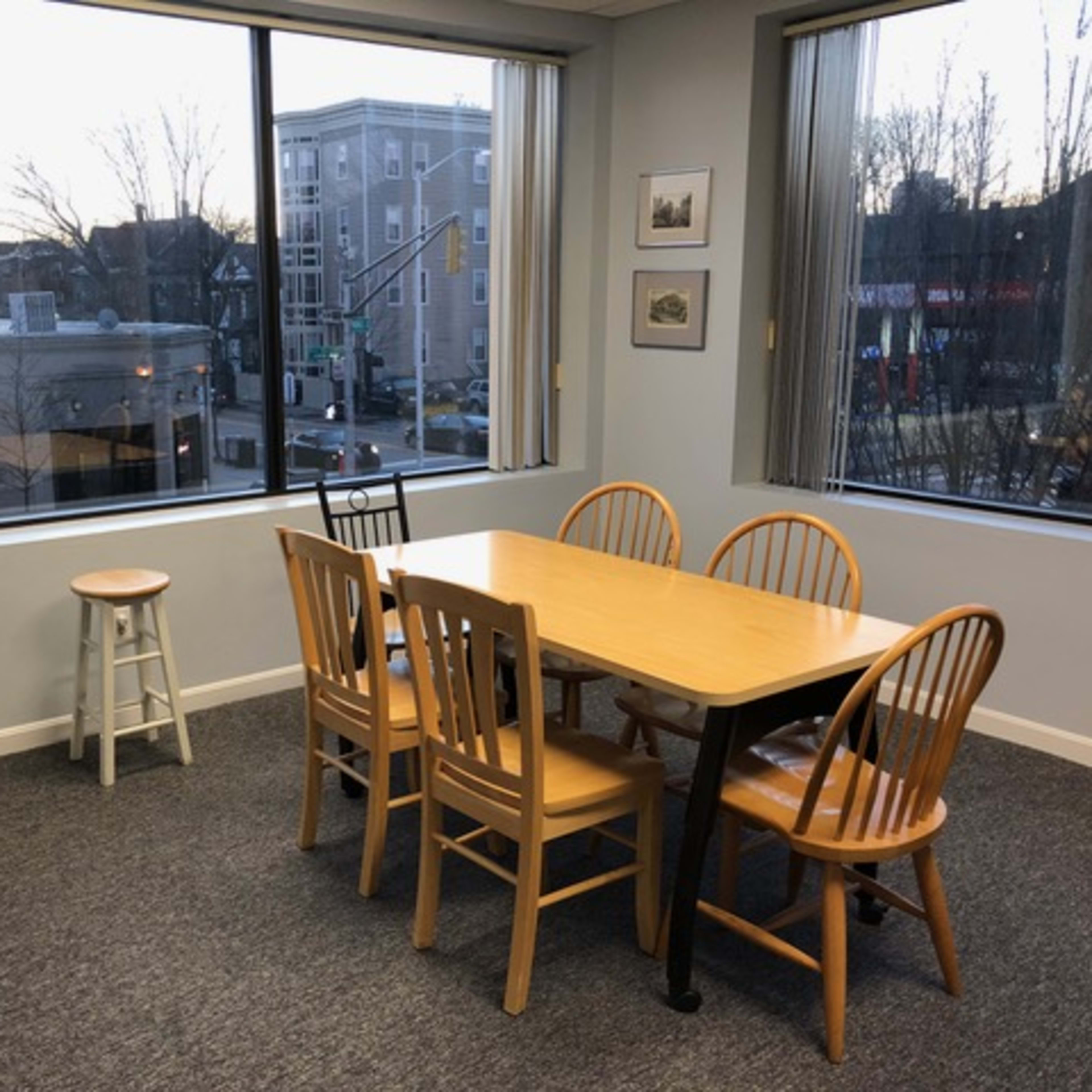 A dining area with a wooden table and six chairs is positioned near large windows overlooking a street scene.