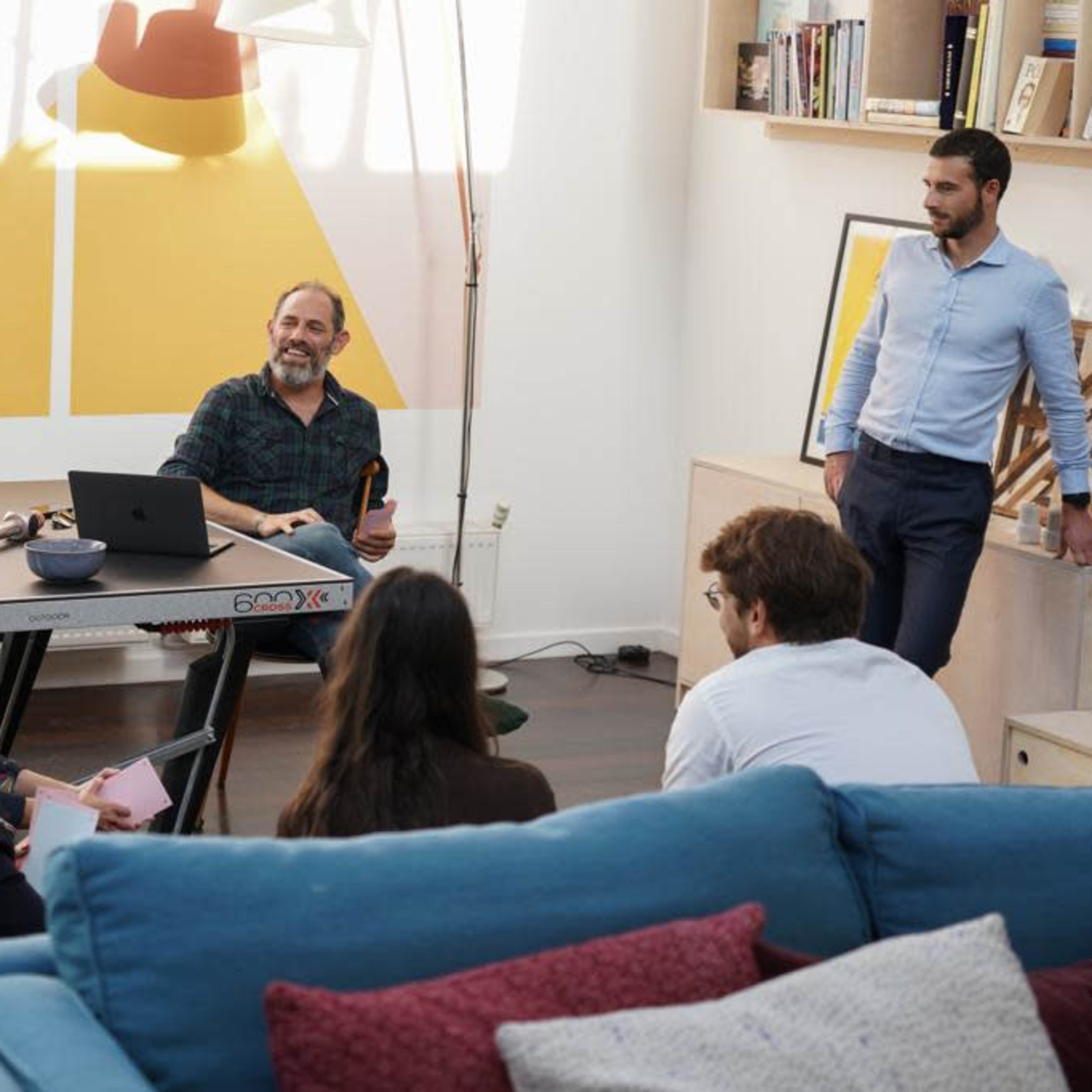 A group of five people sit and stand in a modern living space, engaging in discussion while a man at a desk uses a laptop.