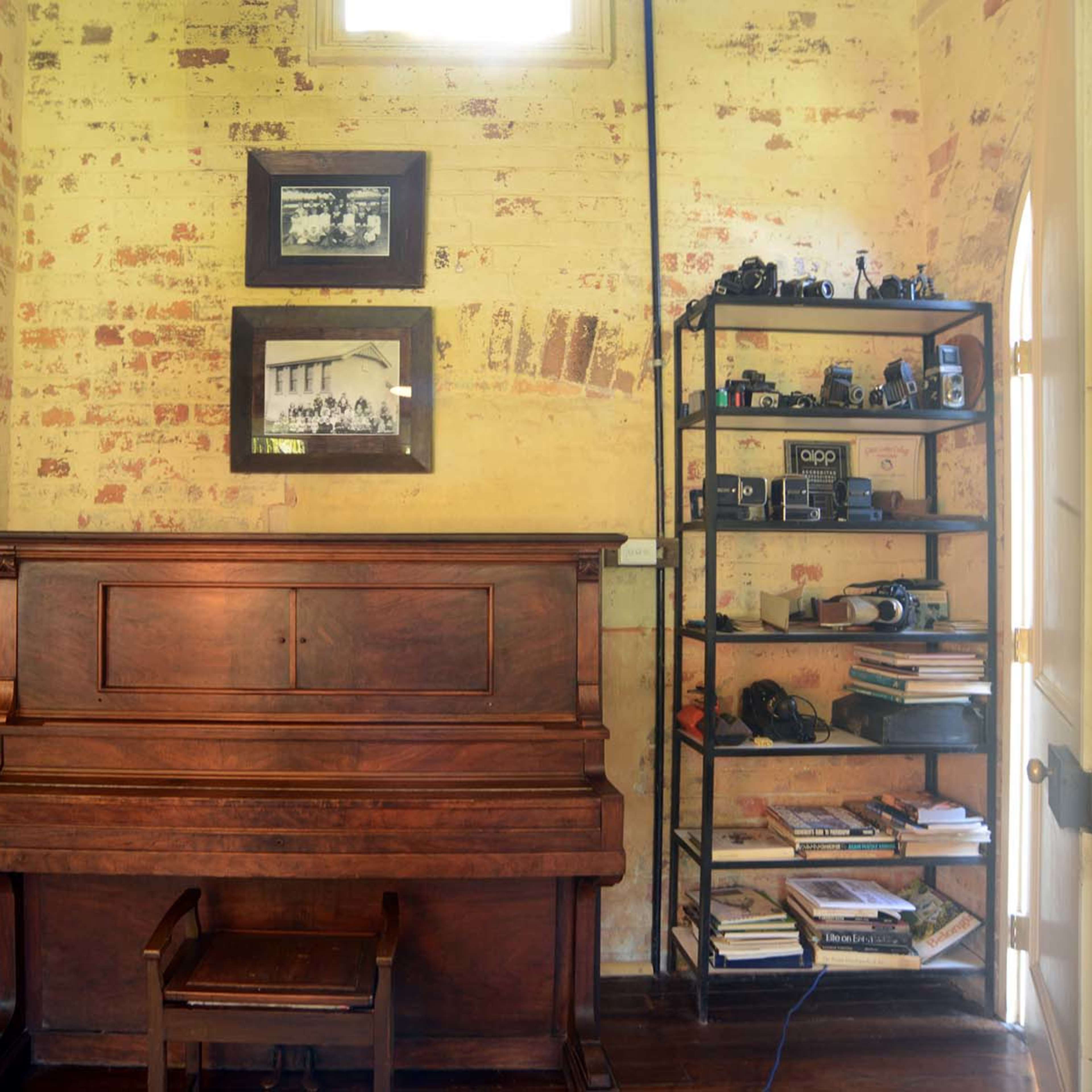 A wooden piano sits against a wall adorned with framed photographs, beside a metal shelf filled with books and cameras.