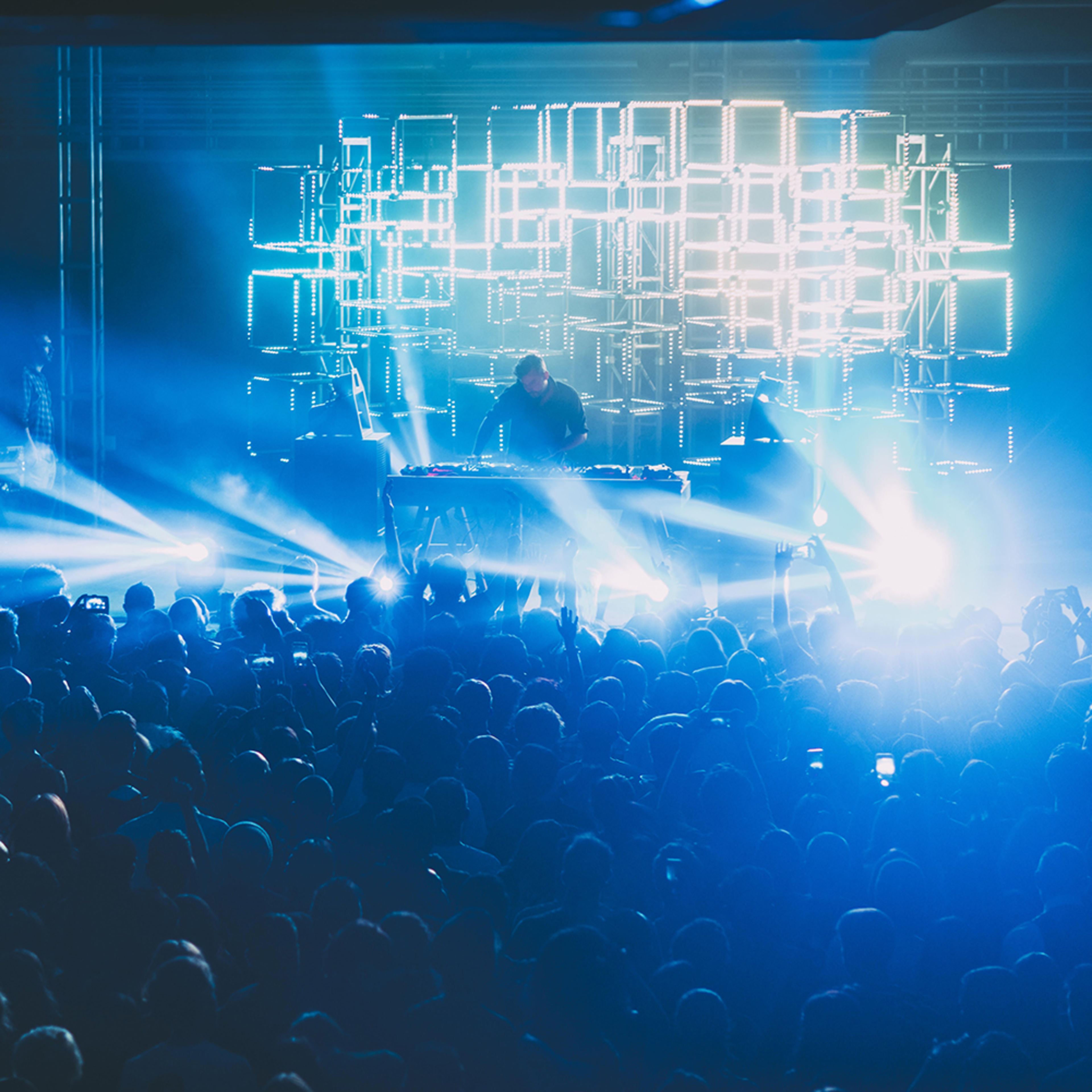 A large crowd gathers in a dimly lit venue as a performer plays electronic music behind a backdrop of illuminated geometric lights.