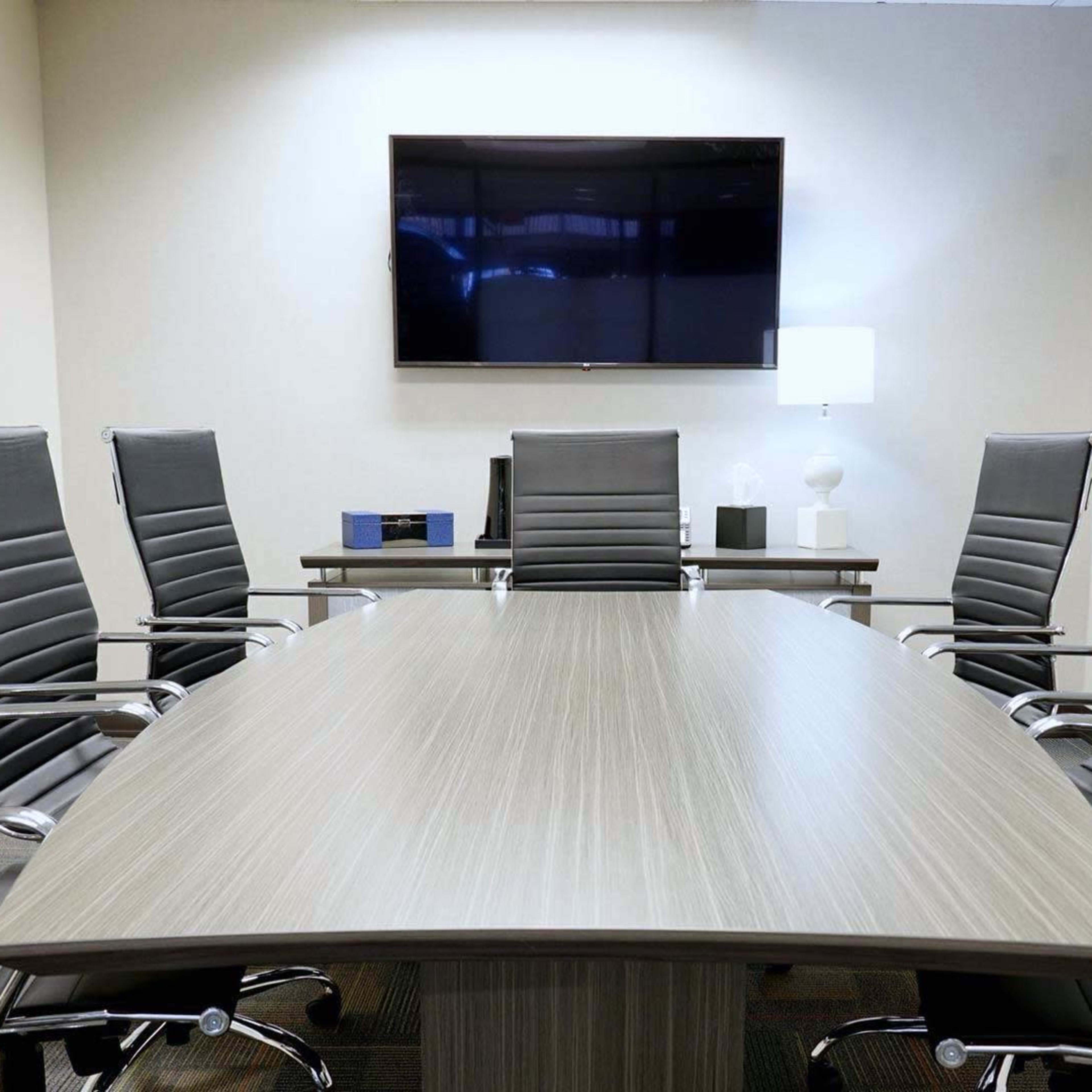 A modern conference room features a large table surrounded by six sleek chairs, with a television mounted on the wall and a lamp on the side.