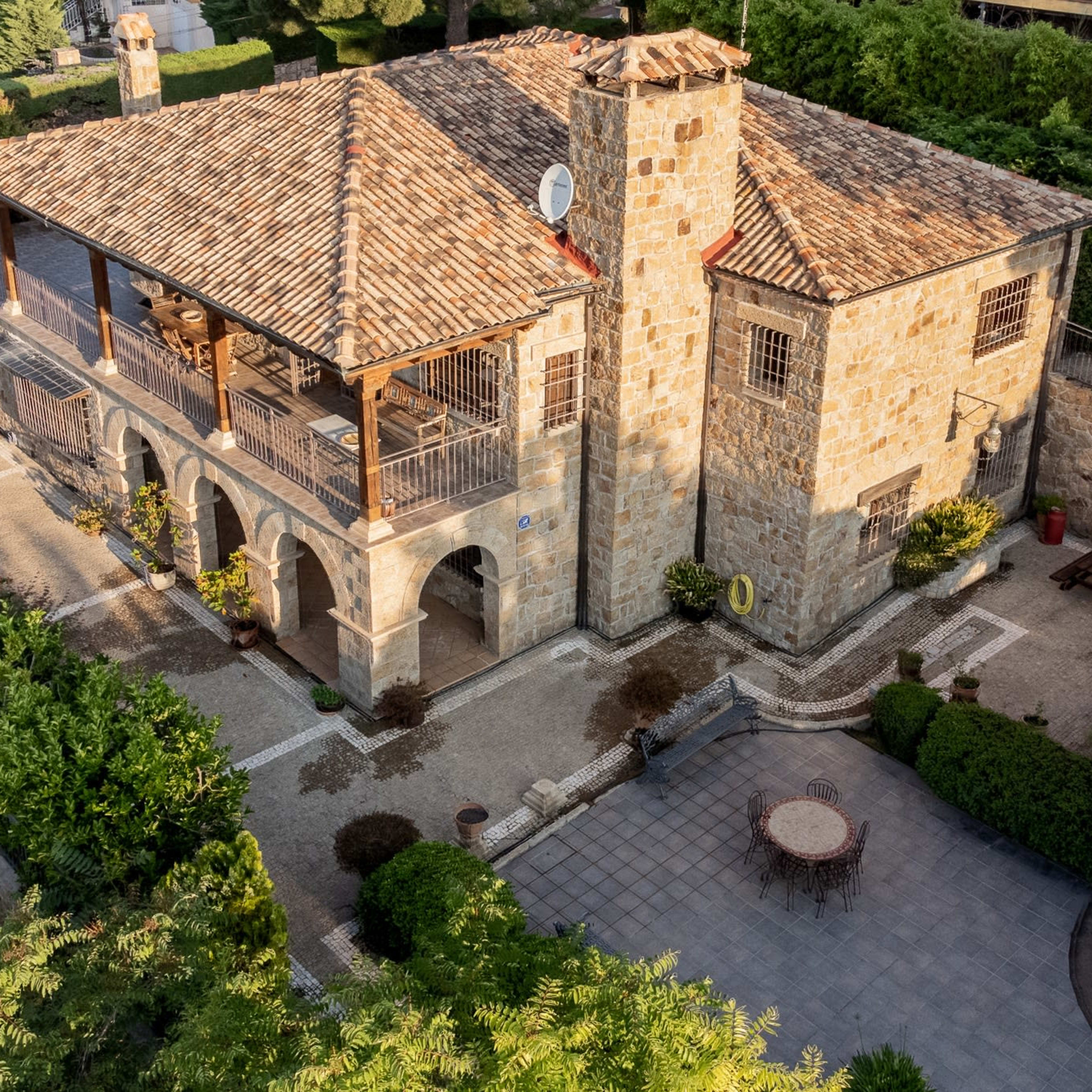 A stone house with a tiled roof, featuring a terrace with arches and surrounded by well-maintained landscaping.