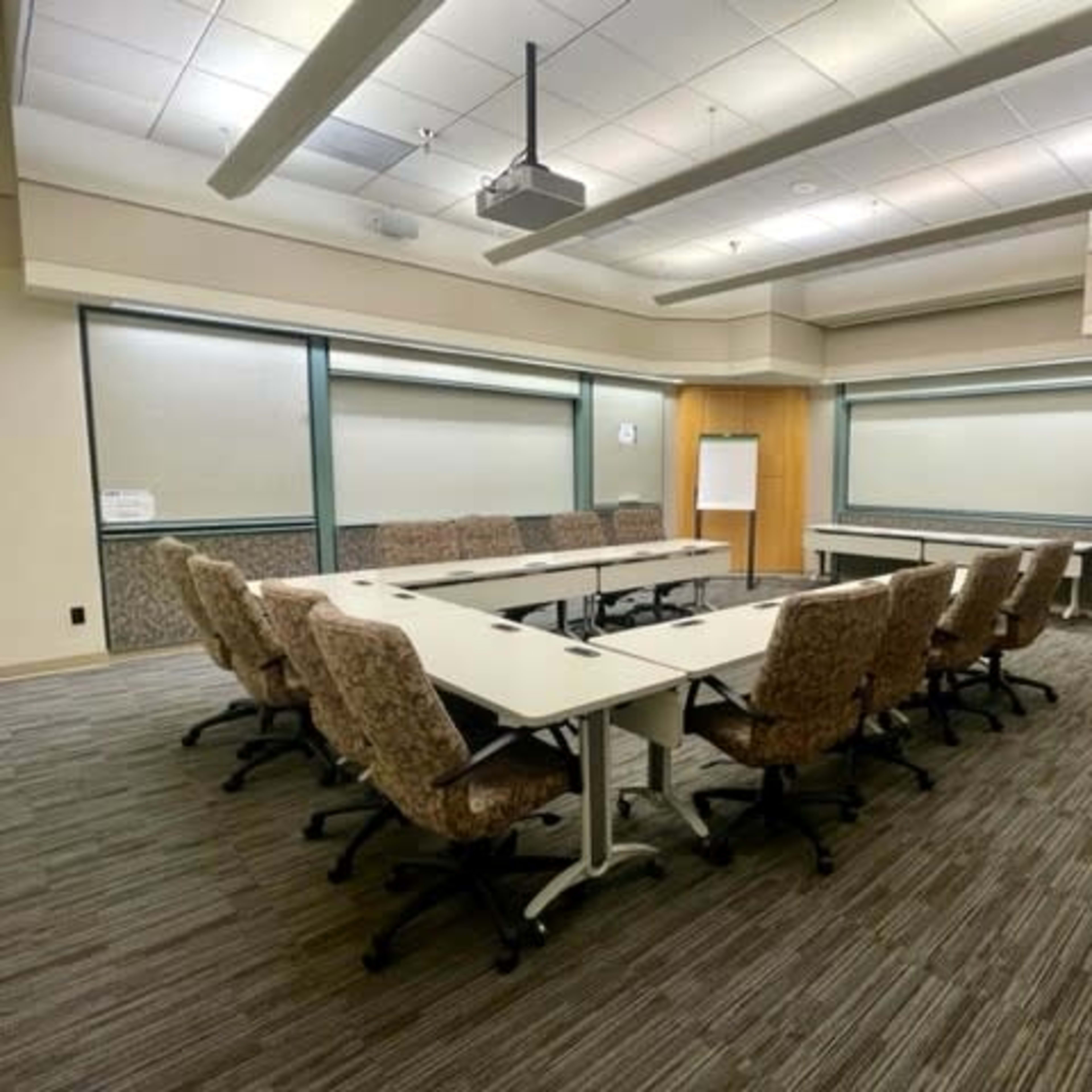 A conference room features a long rectangular table surrounded by upholstered chairs, with a whiteboard and a clock visible in the background.