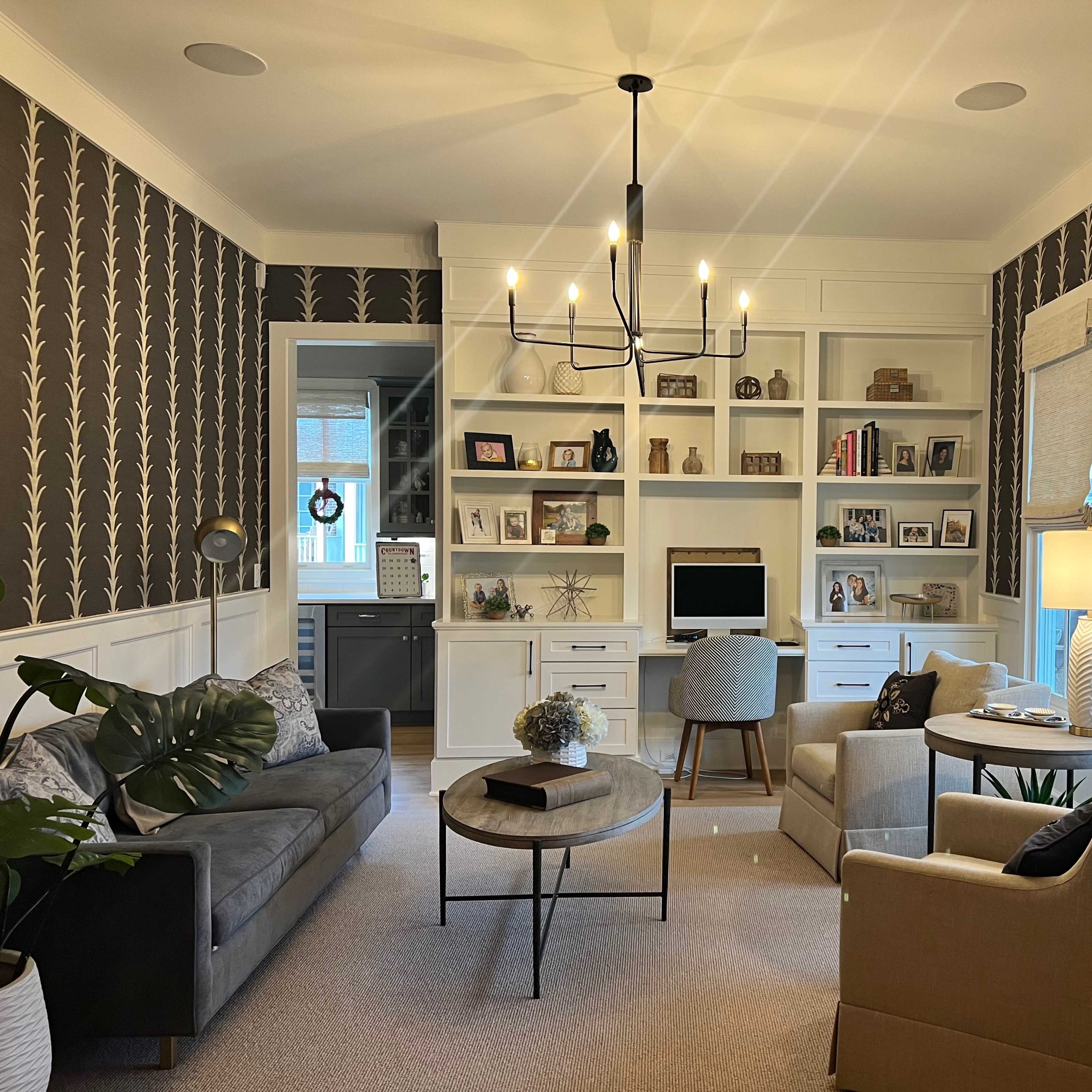 A cozy home office with two sofas, a circular coffee table, a bookshelf filled with decorative items, and a desk with a computer, all set against a patterned wallpaper.