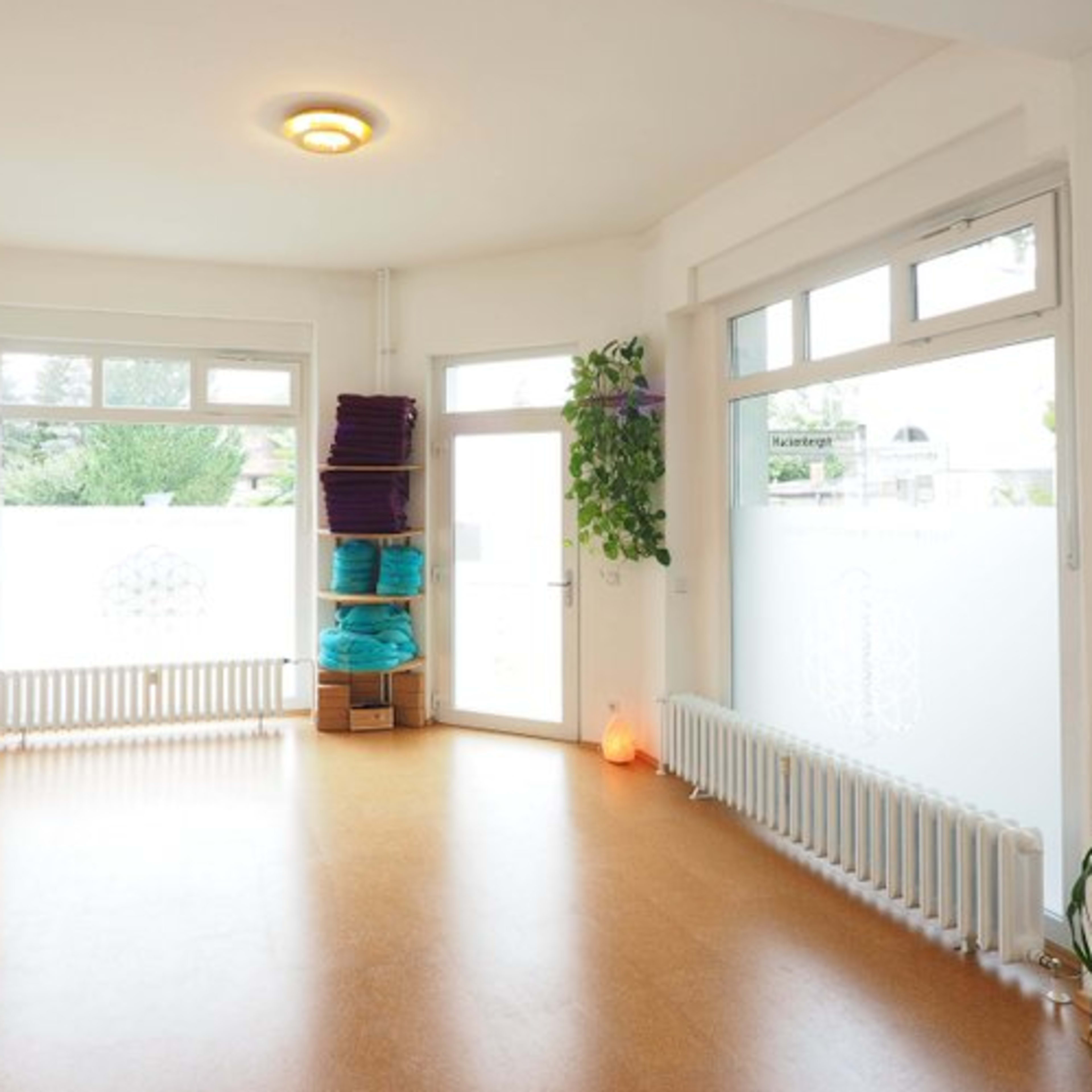 A bright, empty room with large windows, natural light, and plants, featuring a wooden floor and stacked yoga mats.