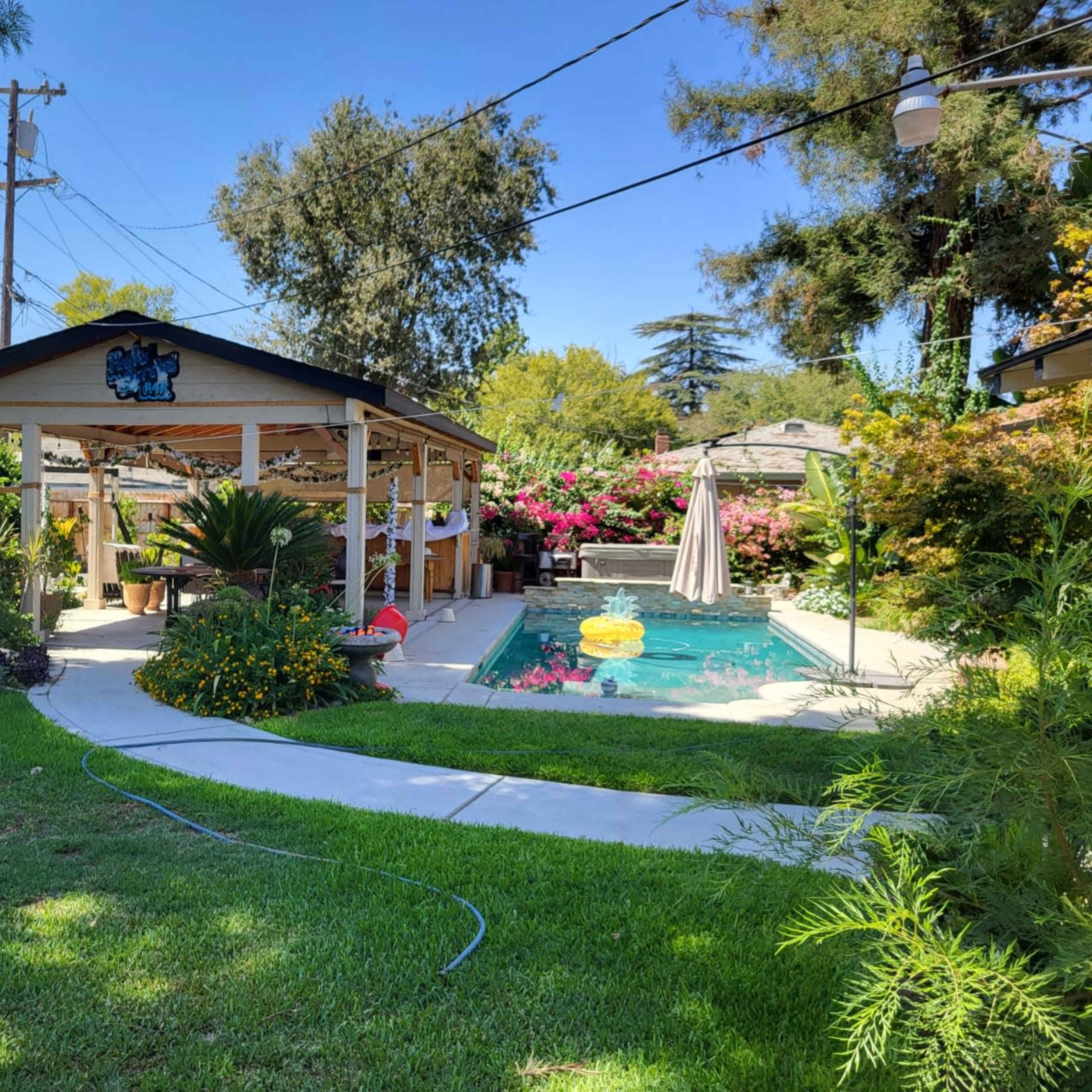 A well-maintained backyard features a swimming pool surrounded by lush plants and a covered patio with seating.