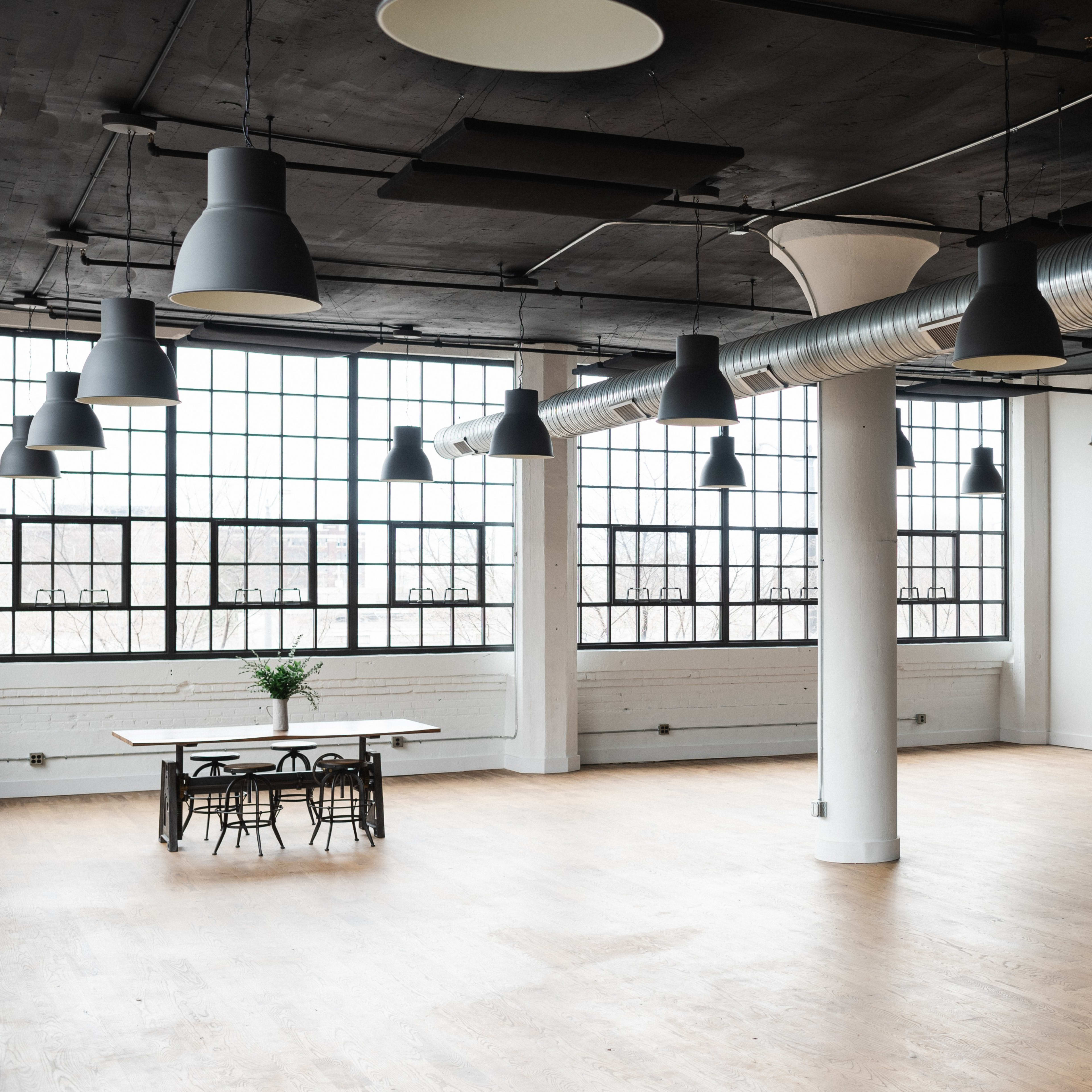 A spacious, modern interior with large windows, exposed ductwork, and a wooden table surrounded by chairs.
