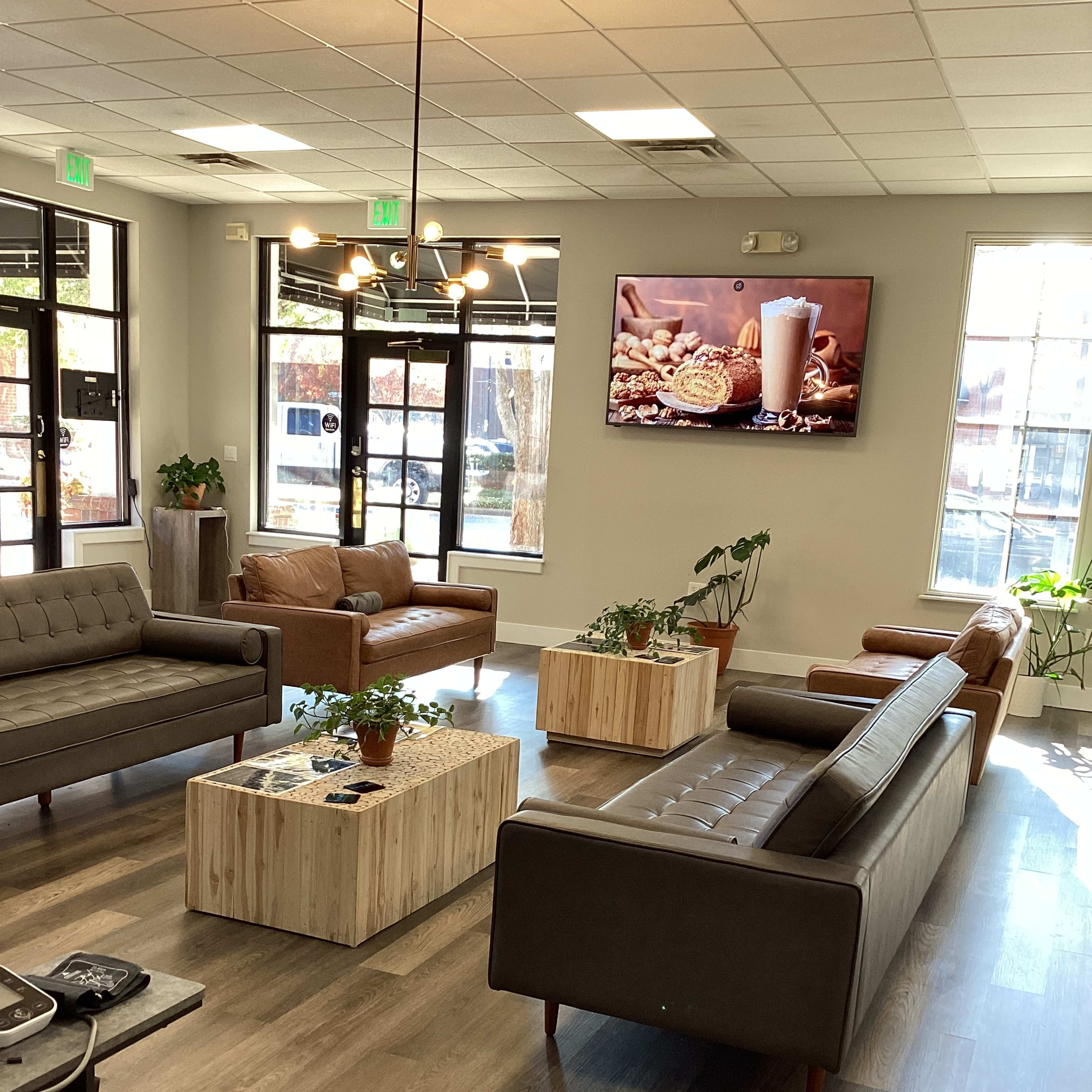 A modern lounge area with several sofas and a coffee table, accompanied by plants and a wall-mounted television displaying food images.