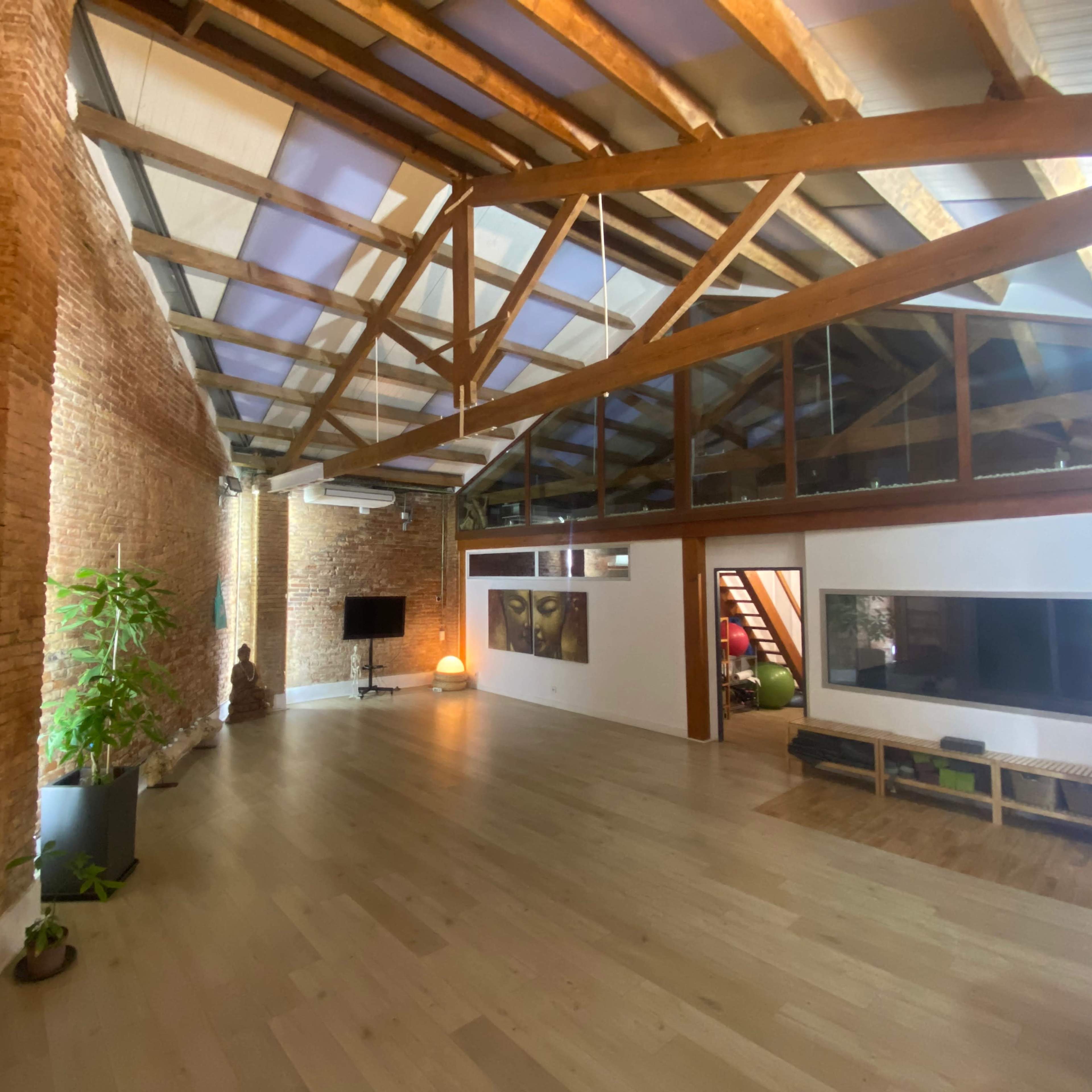 A spacious loft with exposed brick walls, wooden beams, and large windows, featuring a minimalist design and a few plants.