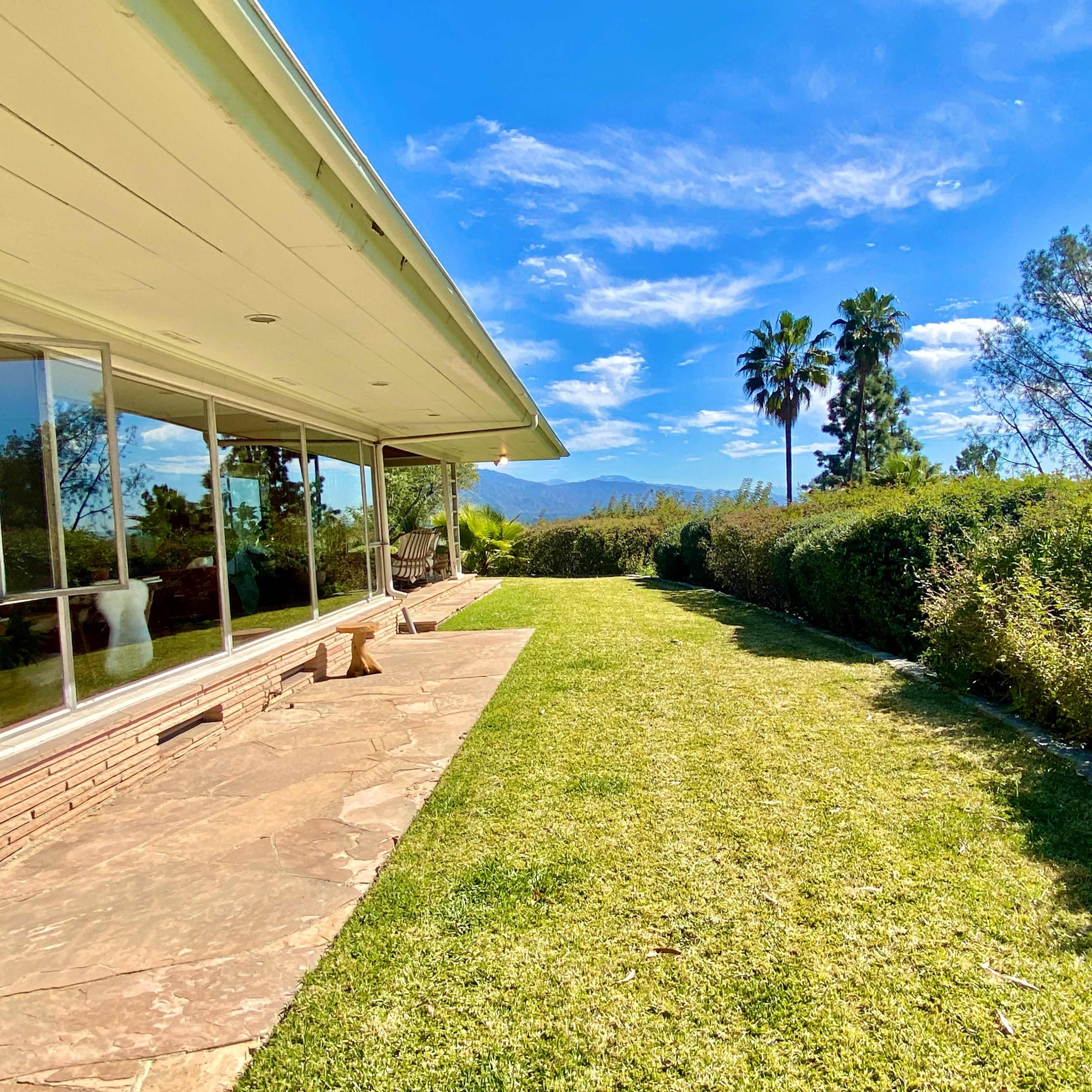 A landscaped yard with a grassy area and a patio adjacent to a modern house, framed by trees and distant mountains under a clear sky.
