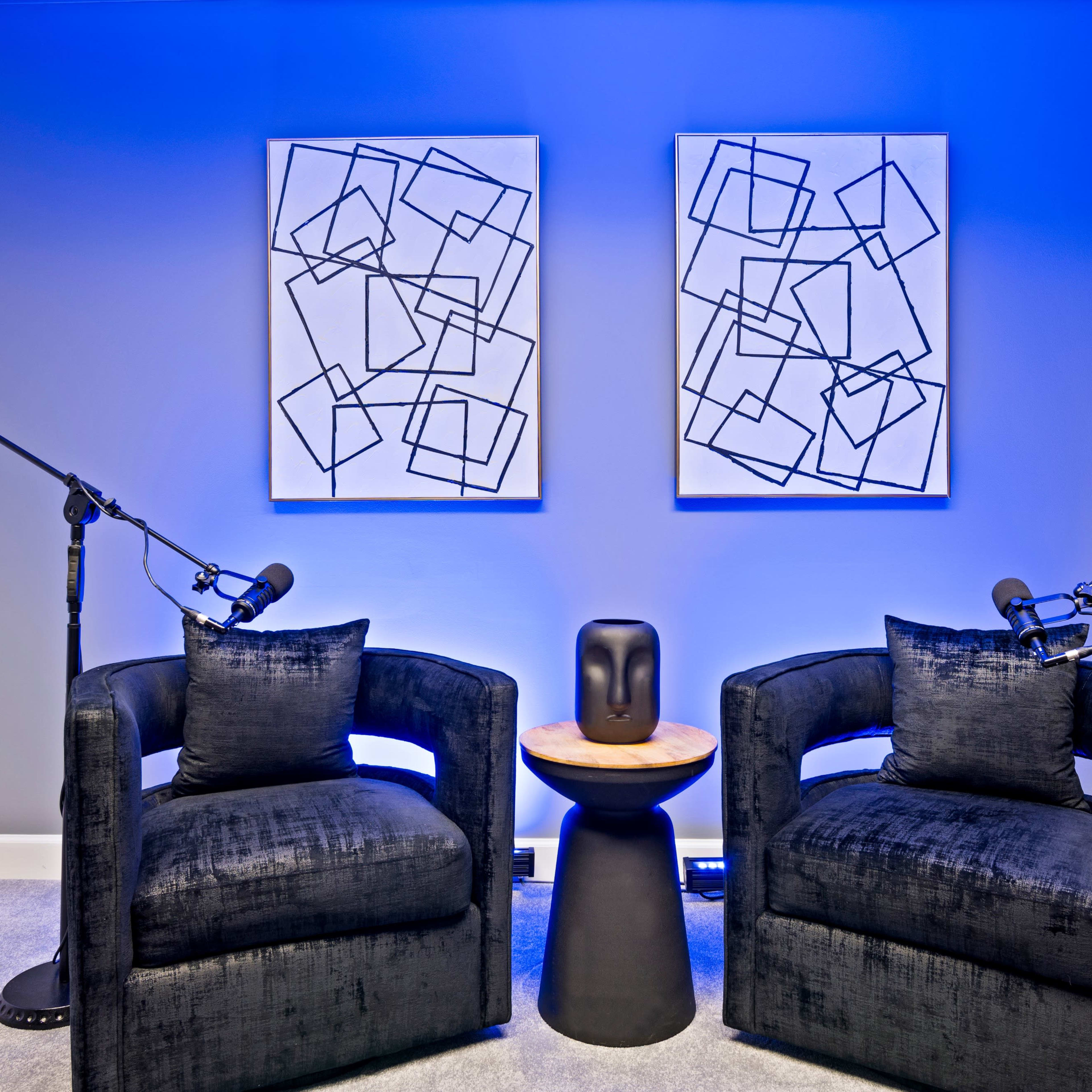 Two black armchairs positioned facing a small round table, with abstract artwork on the walls and microphones set up for recording.