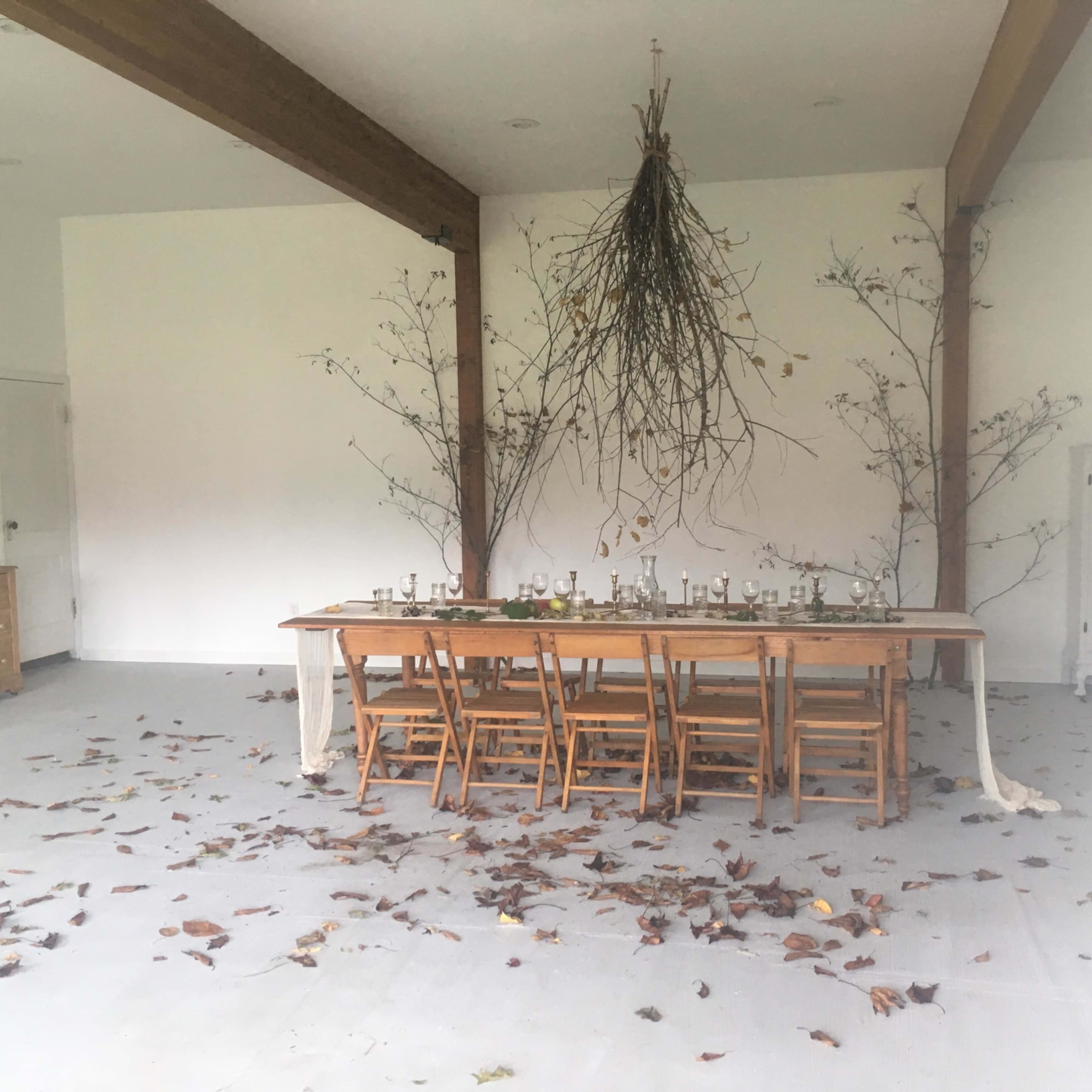 A long wooden table with chairs is set in a room decorated with autumn leaves and branches hanging from the ceiling.